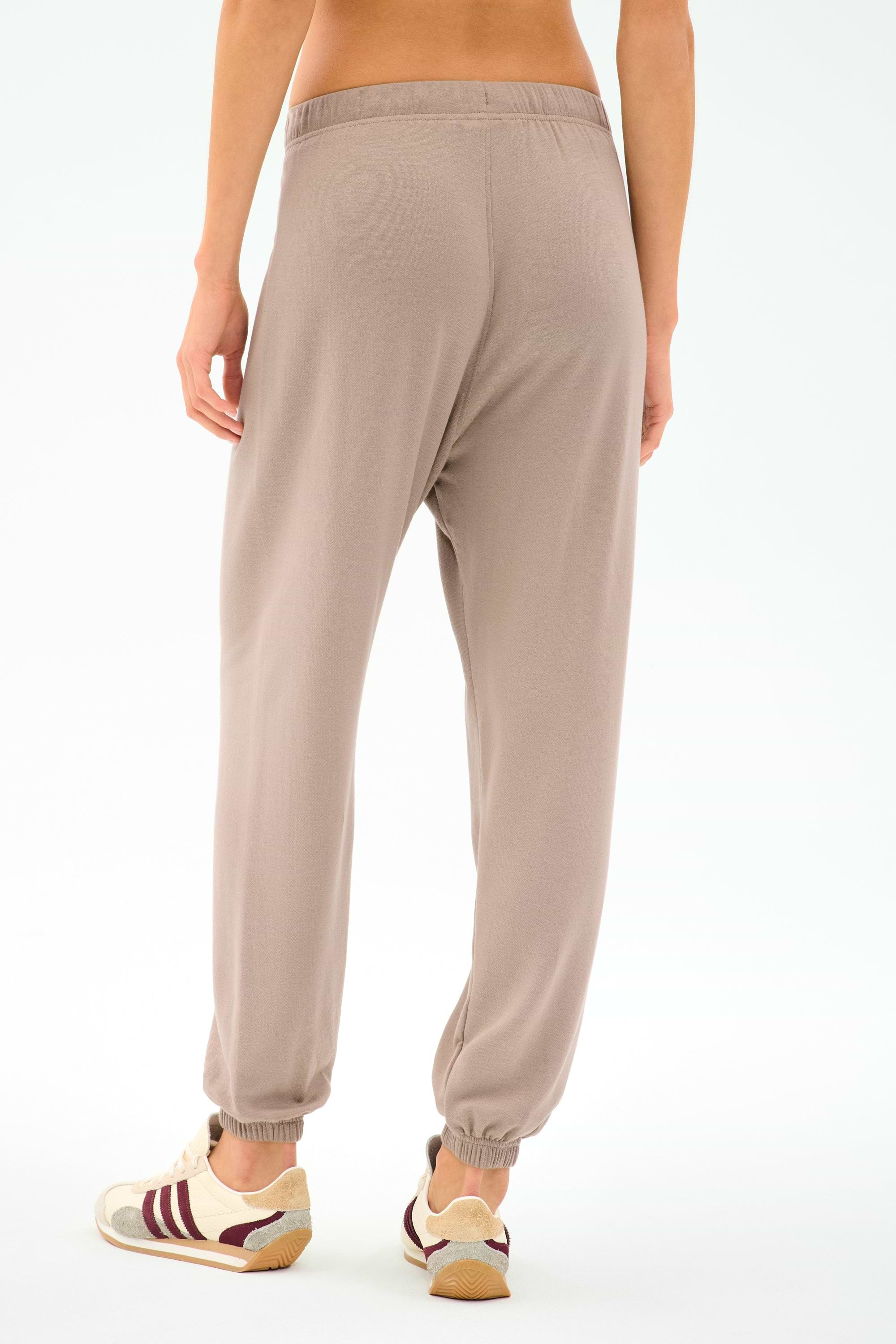 A person stands against a plain white background wearing SPLITS59’s Andie Oversized Fleece Sweatpant in Latte and white sneakers with maroon accents, an ideal look for cool-weather workouts.