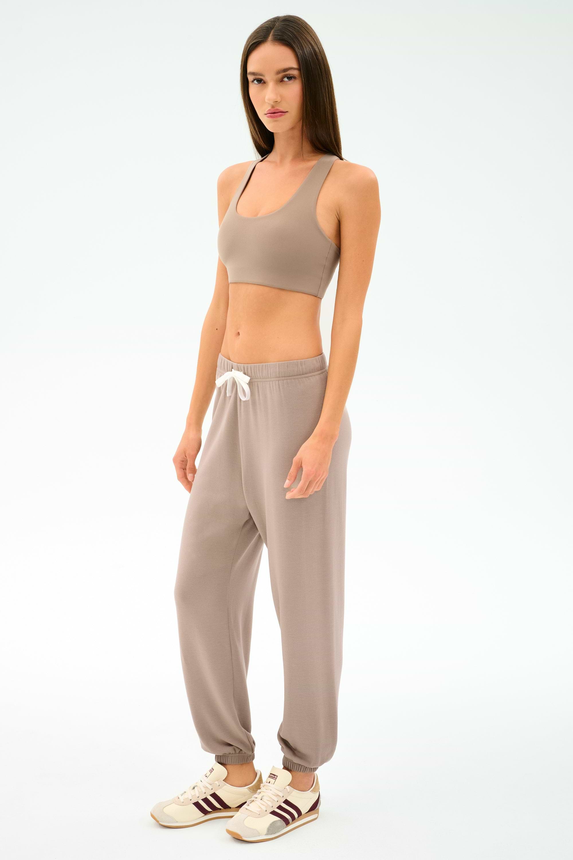 A woman stands against a plain background in a taupe sports bra, SPLITS59 Andie Oversized Fleece Sweatpant - Latte with white drawstring, and white sneakers with brown stripes—ideal for cool weather workouts or warm-ups.