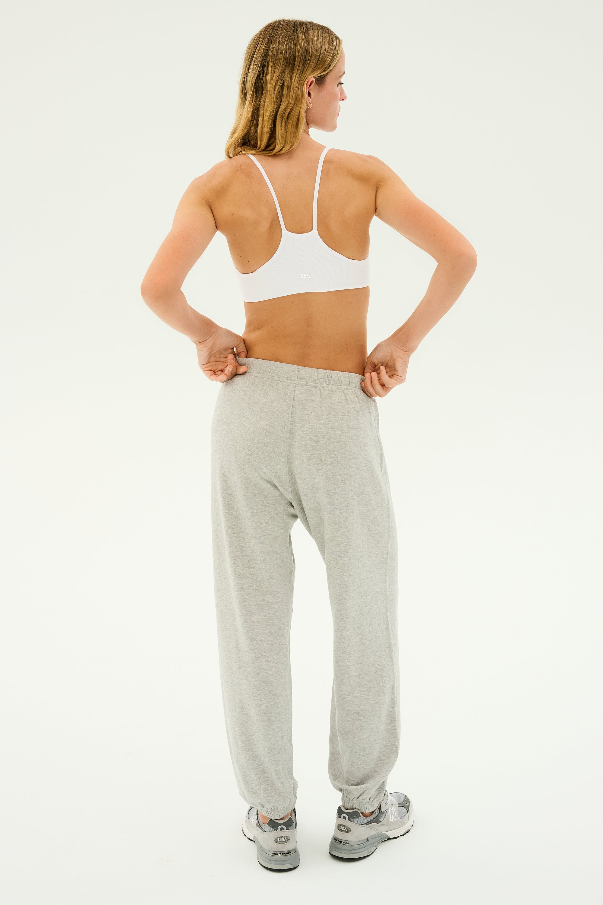 A person in a white sports bra is standing and adjusting the waistband of their Andie Oversized Fleece Sweatpant in Heather Grey by SPLITS59. The person is facing away from the camera with their head turned slightly to the side, making it perfect for cool-weather workouts.