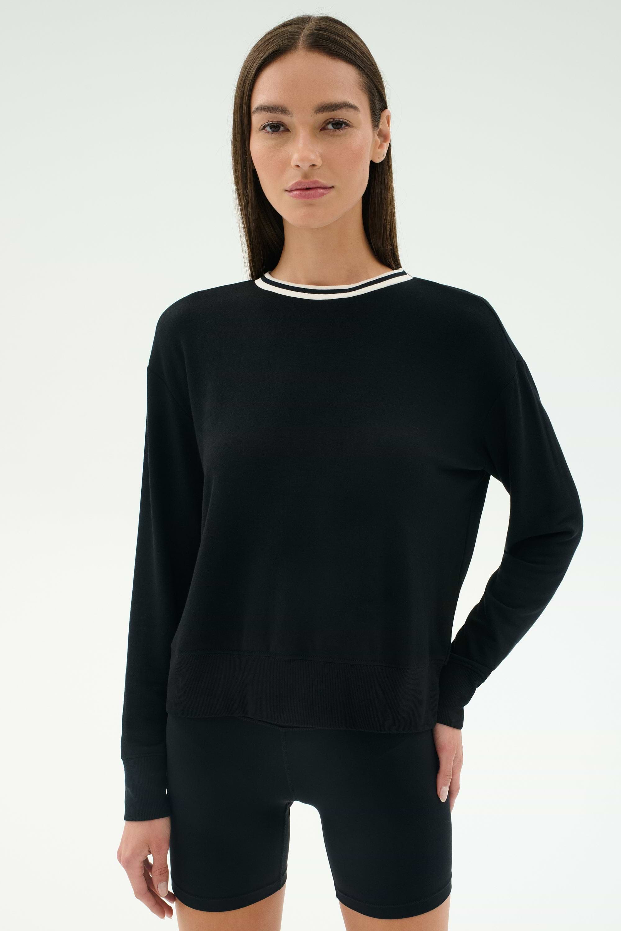 A woman faces forward in a SPLITS59 Sonja Fleece Sweatshirt W/ Stripe in black, featuring a white-trimmed collar, paired with black shorts against a plain light background.