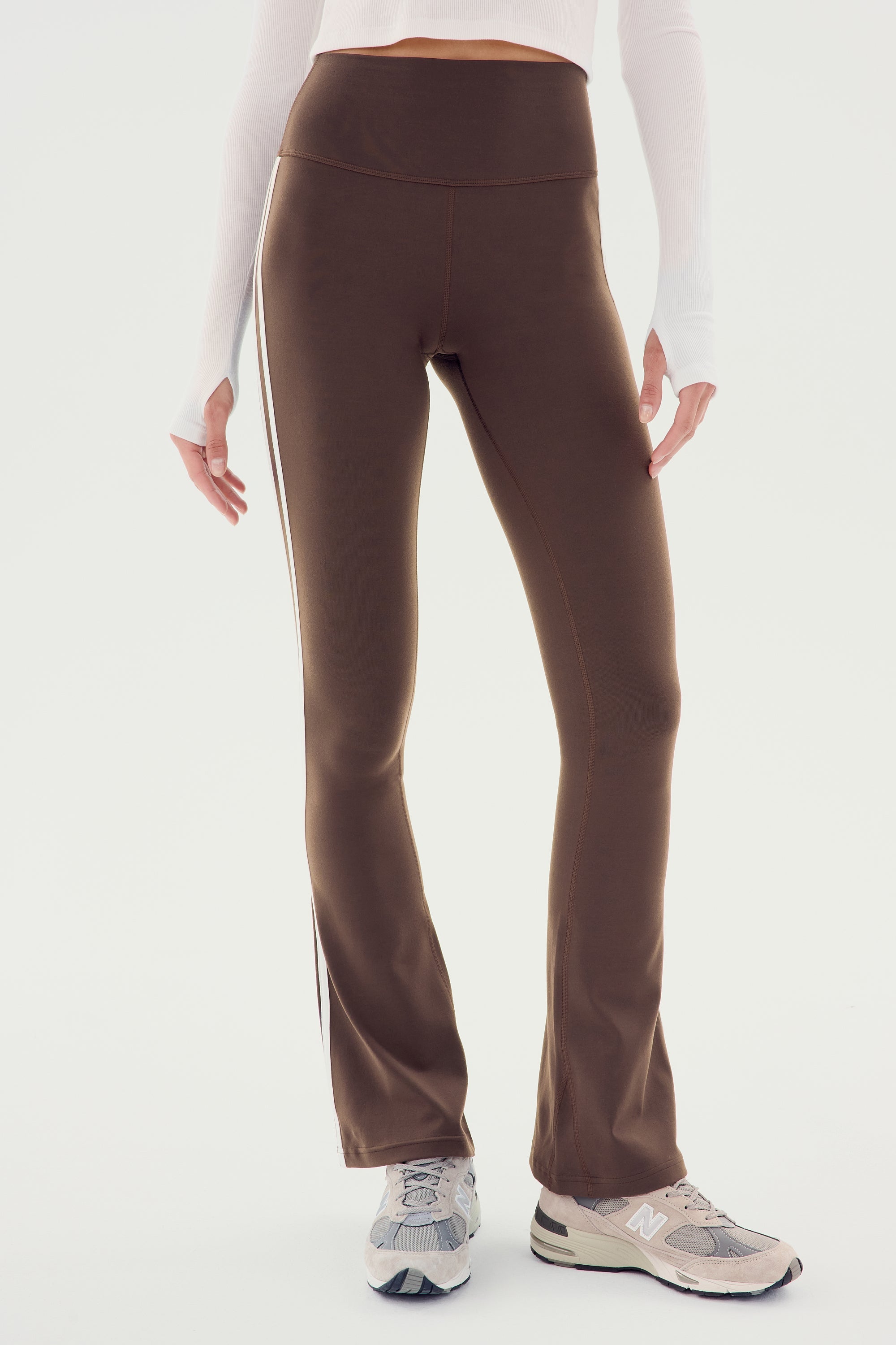A person wearing SPLITS59 Raquel High Waist Flared Legging in Dark Chocolate with White side stripes, made from 4-way stretch supplex fabric, and running shoes is standing against a white background. Only the lower torso and legs are visible.