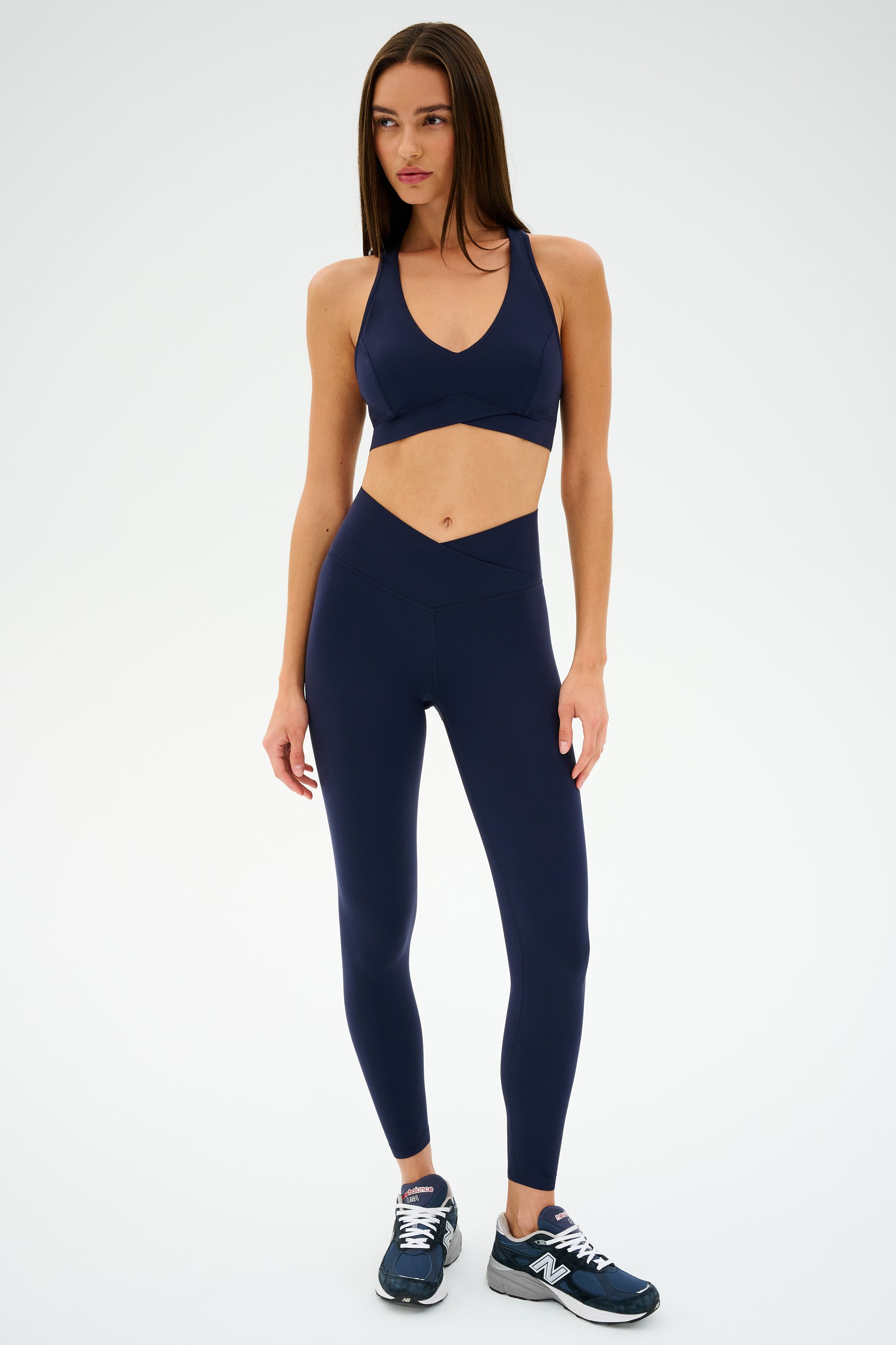 A person in an Indigo Mia Rigor Bra by SPLITS59, paired with leggings designed for multi-sport use, and dark athletic shoes stands against a light background.