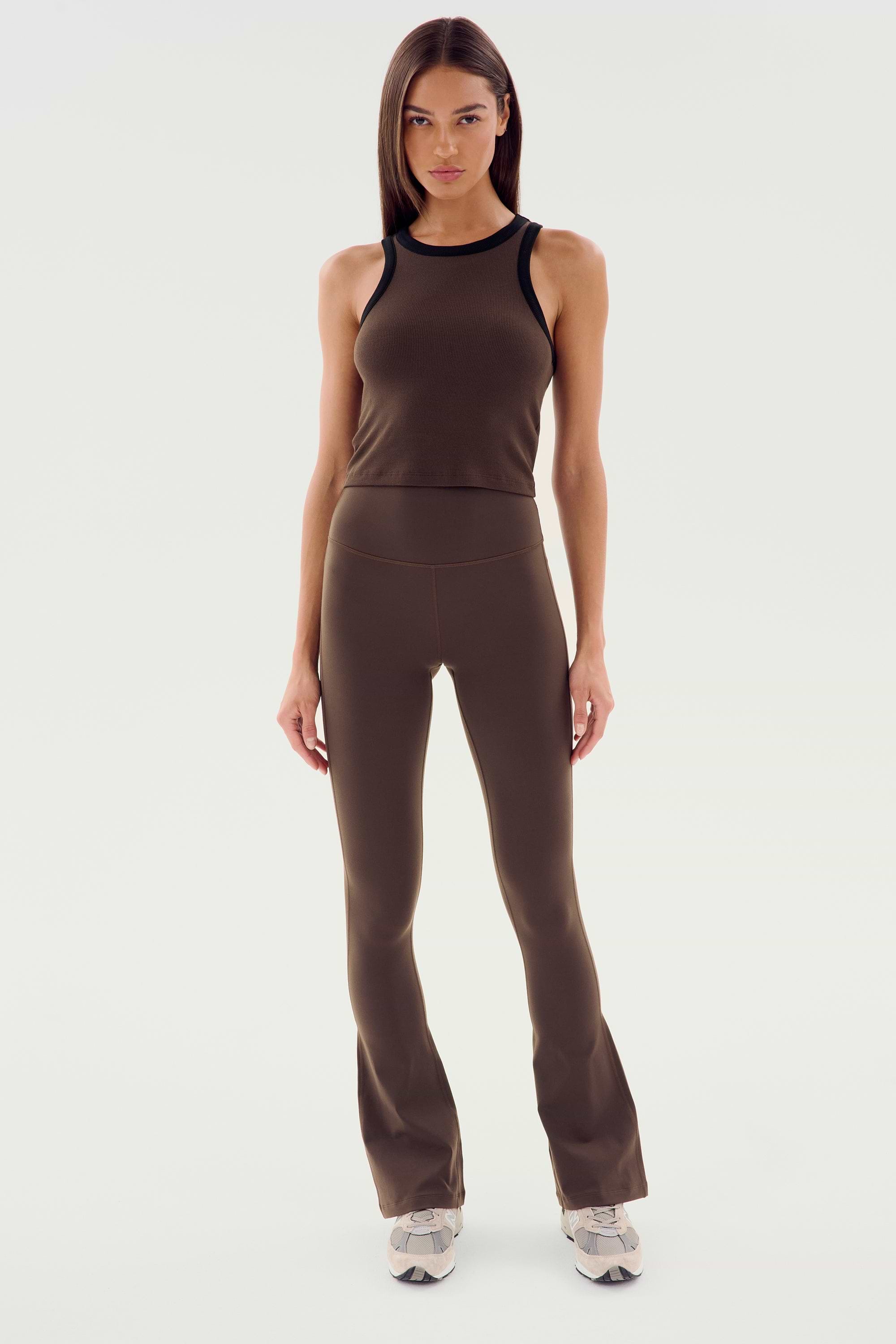 A woman stands in front of a plain background, wearing a sleeveless brown top, Raquel High Waist Flared Legging in Dark Chocolate by SPLITS59, and white sneakers. She has straight brown hair and a neutral expression.