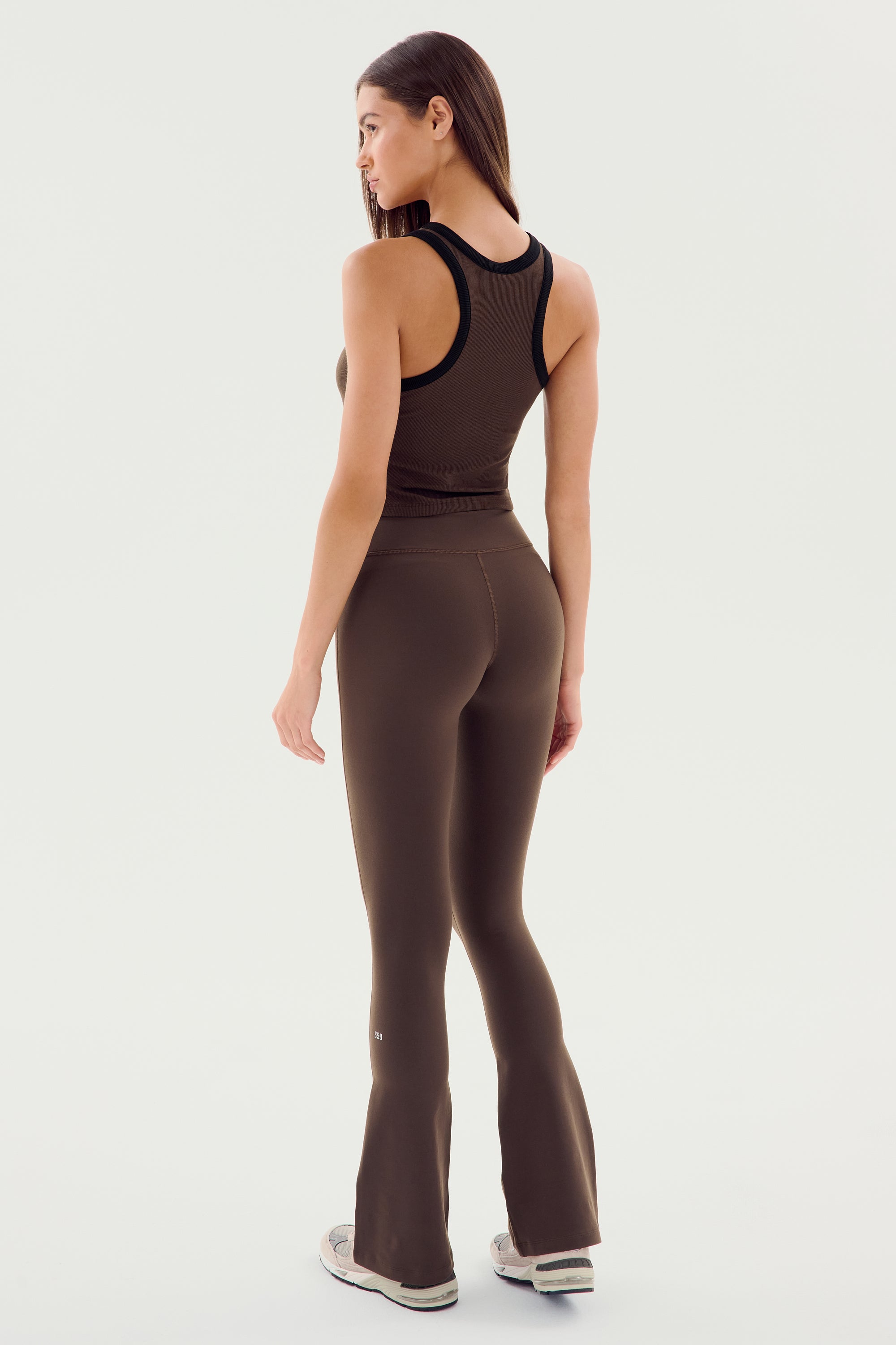 Woman facing sideways, wearing a SPLITS59 Raquel High Waist Flared Legging in Dark Chocolate and a matching sleeveless top with 4-way stretch, paired with white sneakers, on a plain light background.