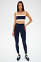 A woman stands against a white background wearing the SPLITS59 Monah Rigor Bra in Indigo/Creme, paired with high-waisted nylon leggings and navy sneakers.