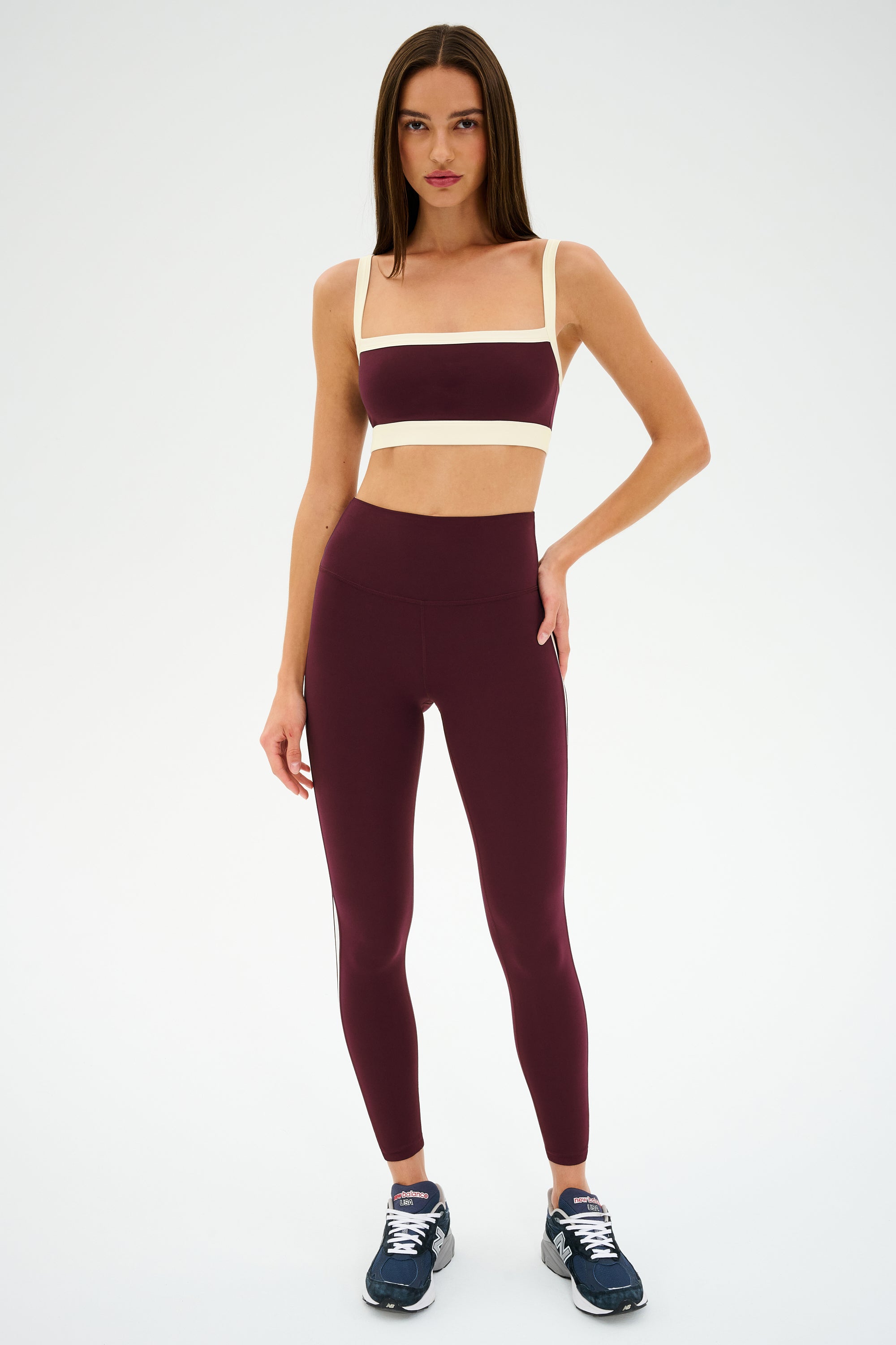 A woman stands against a plain background wearing the SPLITS59 Clare High Waist Rigor 7/8 leggings in Dark Cherry/Creme, paired with a matching sports bra, both crafted from a soft nylon-spandex blend and blue sneakers.