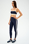 A woman wears the SPLITS59 Monah Rigor Bra in Indigo/Creme with matching leggings and navy running shoes, standing against a plain white background.