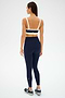 A woman stands against a plain backdrop, shown from the back, wearing the SPLITS59 Monah Rigor Bra in Indigo/Creme and matching leggings with white trim, paired with black athletic shoes.