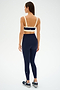 A woman stands against a plain backdrop, shown from the back, wearing the SPLITS59 Monah Rigor Bra in Indigo/Creme and matching leggings with white trim, paired with black athletic shoes.