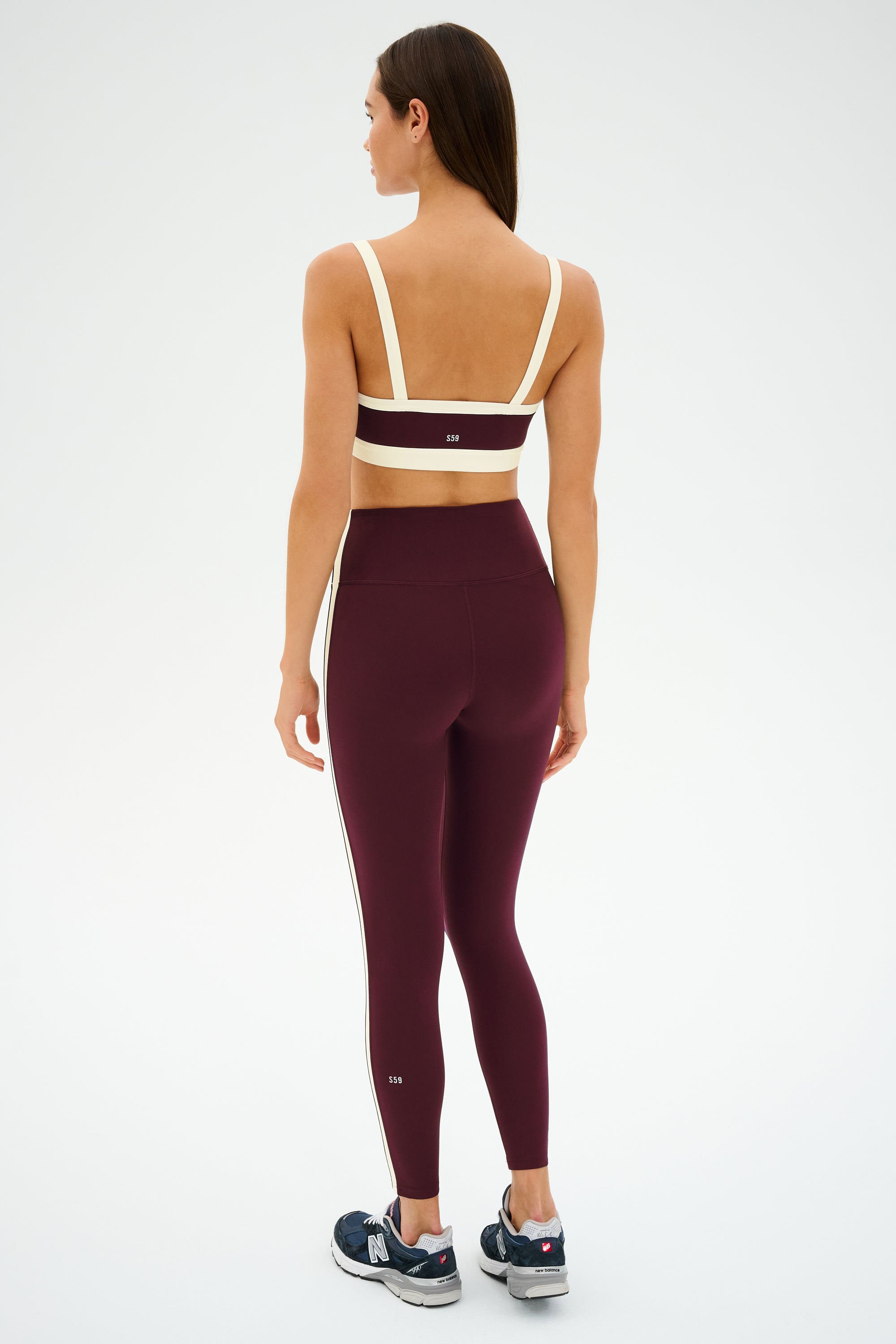 A woman stands facing away, wearing the SPLITS59 Clare High Waist Rigor 7/8 leggings in Dark Cherry/Creme, a beige sports bra, and black athletic shoes against a simple light background.