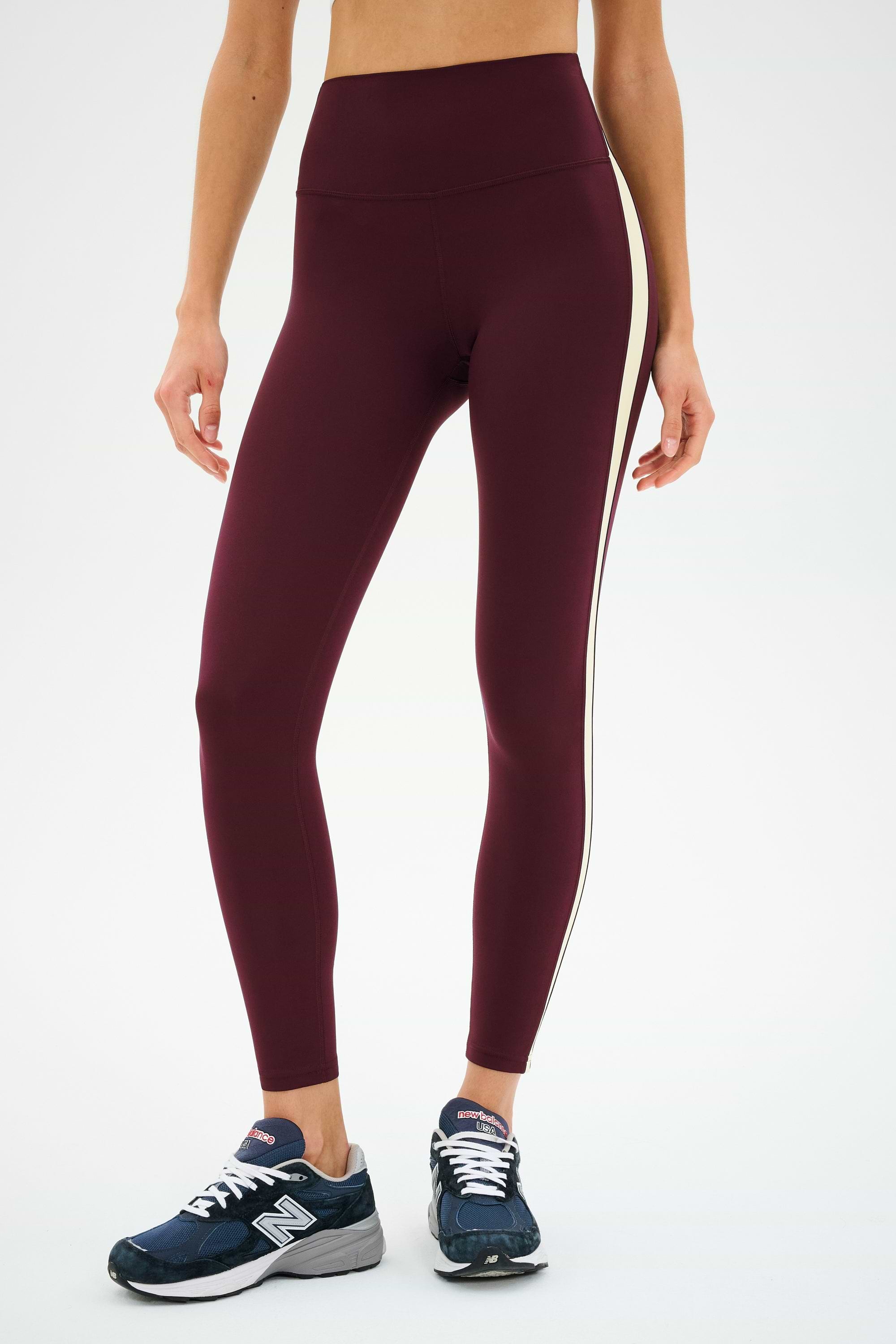 A person wearing SPLITS59 Clare High Waist Rigor 7/8 leggings in Dark Cherry/Creme with a white side stripe and navy New Balance sneakers, standing against a plain white background.