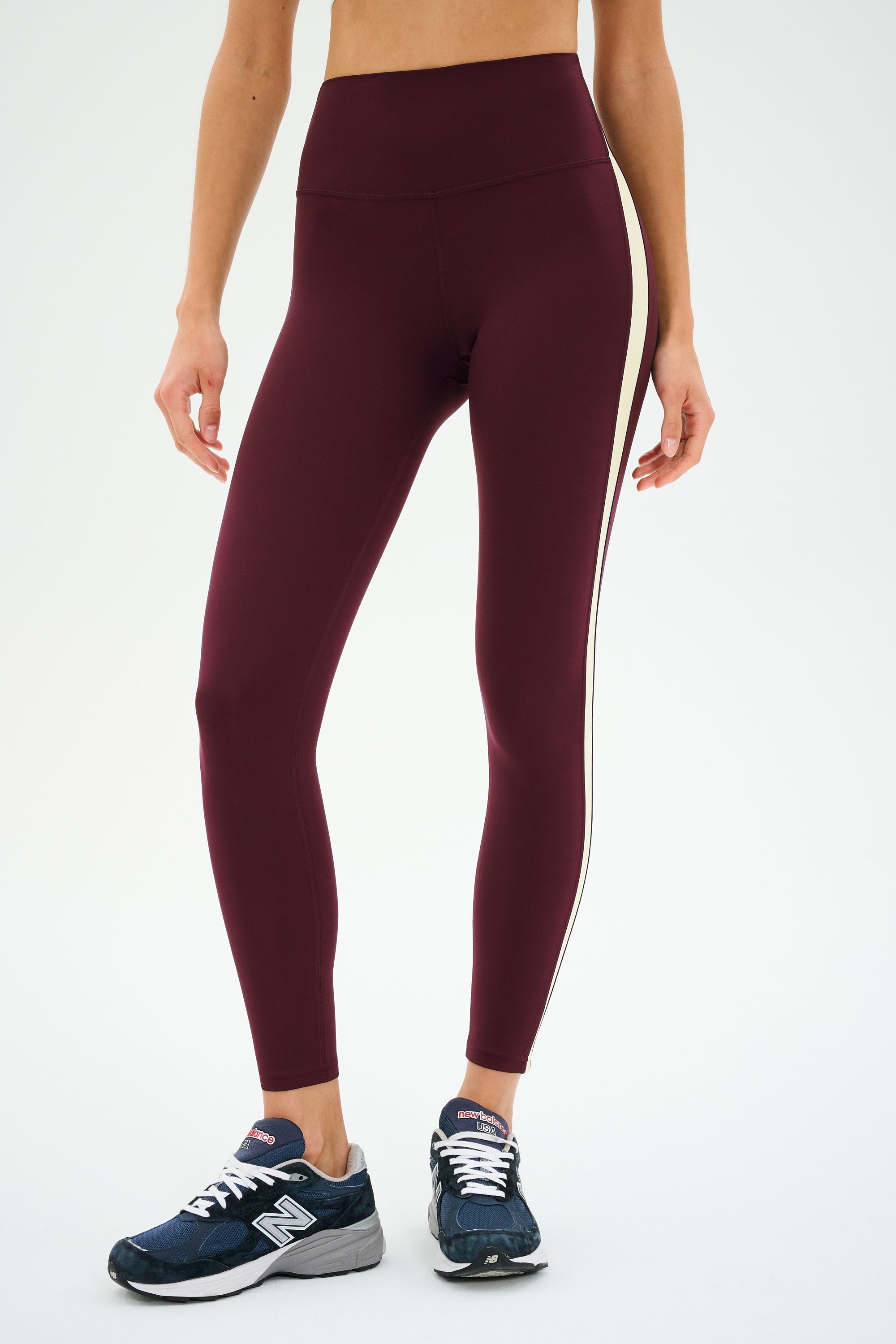 A person wearing SPLITS59 Clare High Waist Rigor 7/8 leggings in Dark Cherry/Creme with a white side stripe and navy New Balance sneakers, standing against a plain white background.