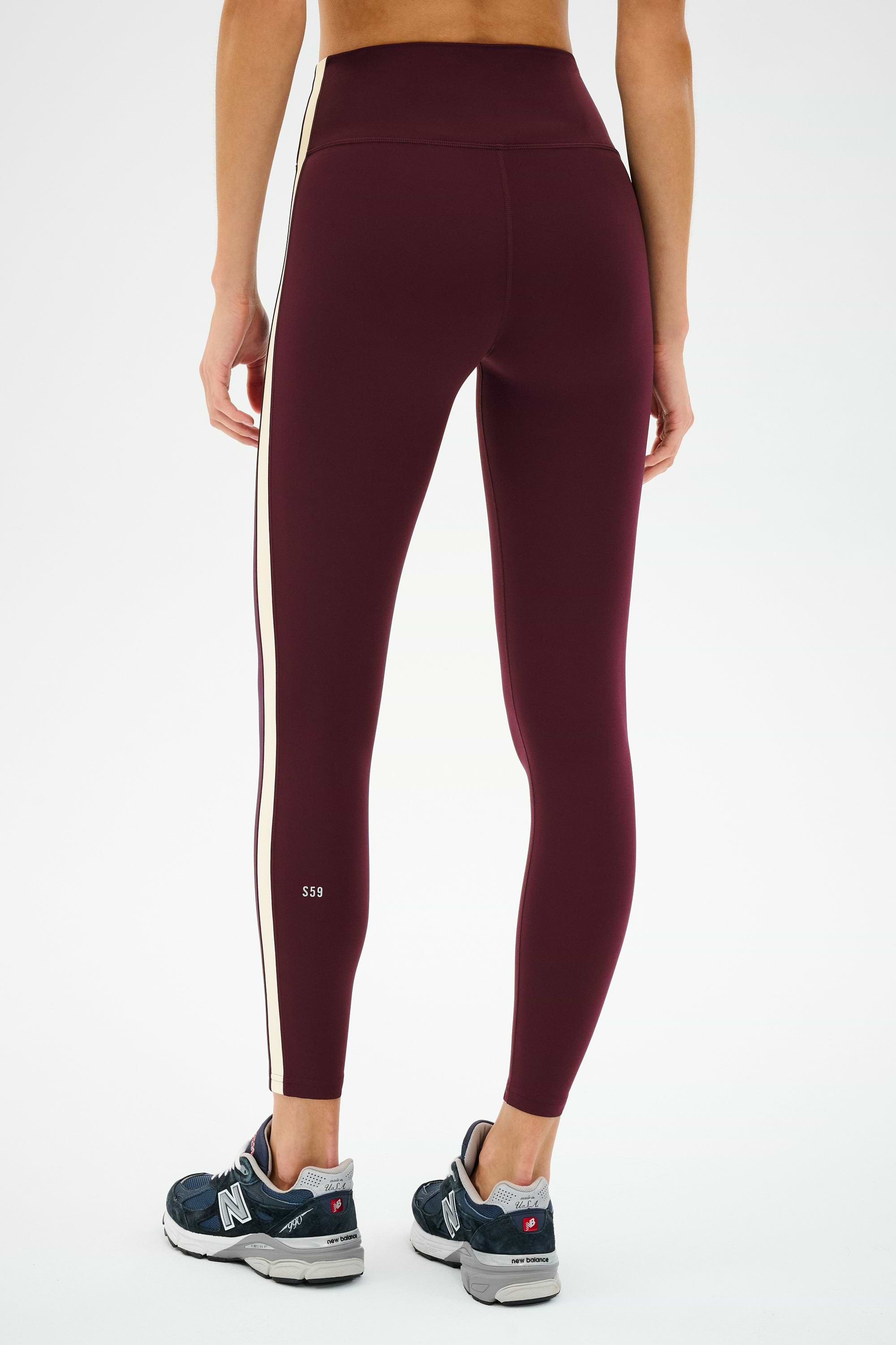 A person wears SPLITS59 Clare High Waist Rigor 7/8 leggings in Dark Cherry/Creme, accented with cream side stripes, paired with gray New Balance sneakers, standing against a plain white background.