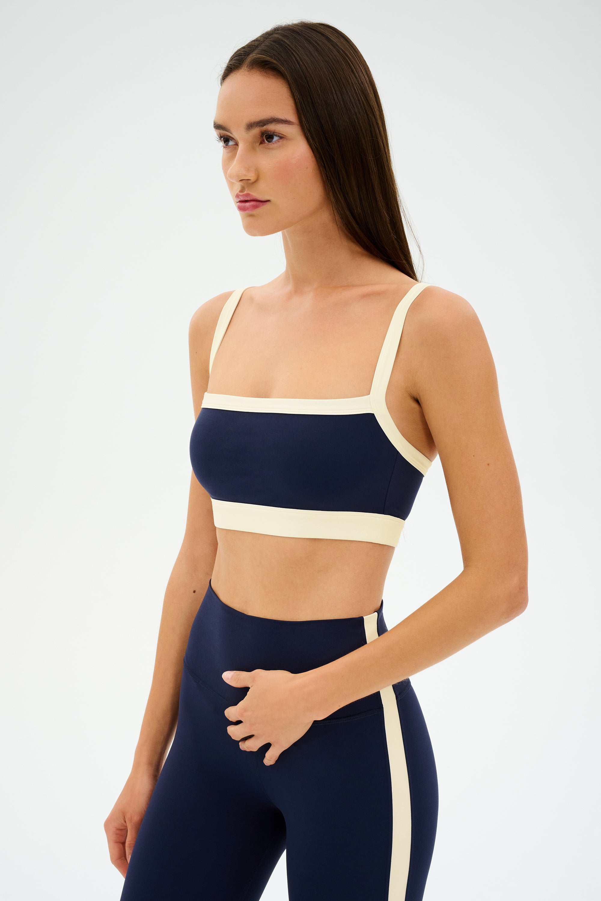 A woman wears the SPLITS59 Monah Rigor Bra in Indigo/Creme, featuring navy blue spandex with beige trim, standing against a plain white background.