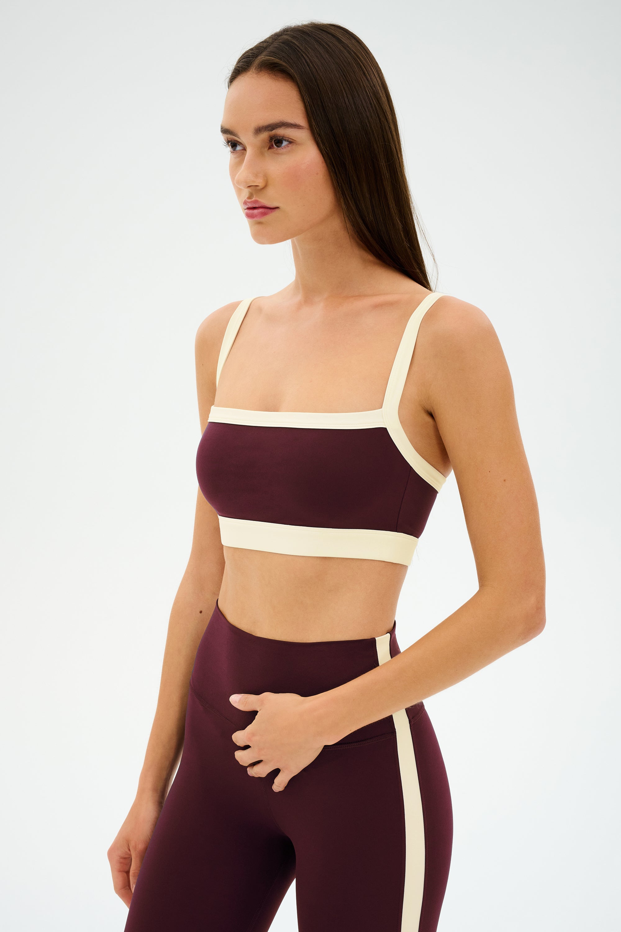 A woman models the SPLITS59 Monah Rigor Bra in Dark Cherry/Creme, a soft nylon-spandex workout set, standing against a plain white background and glancing to the side.