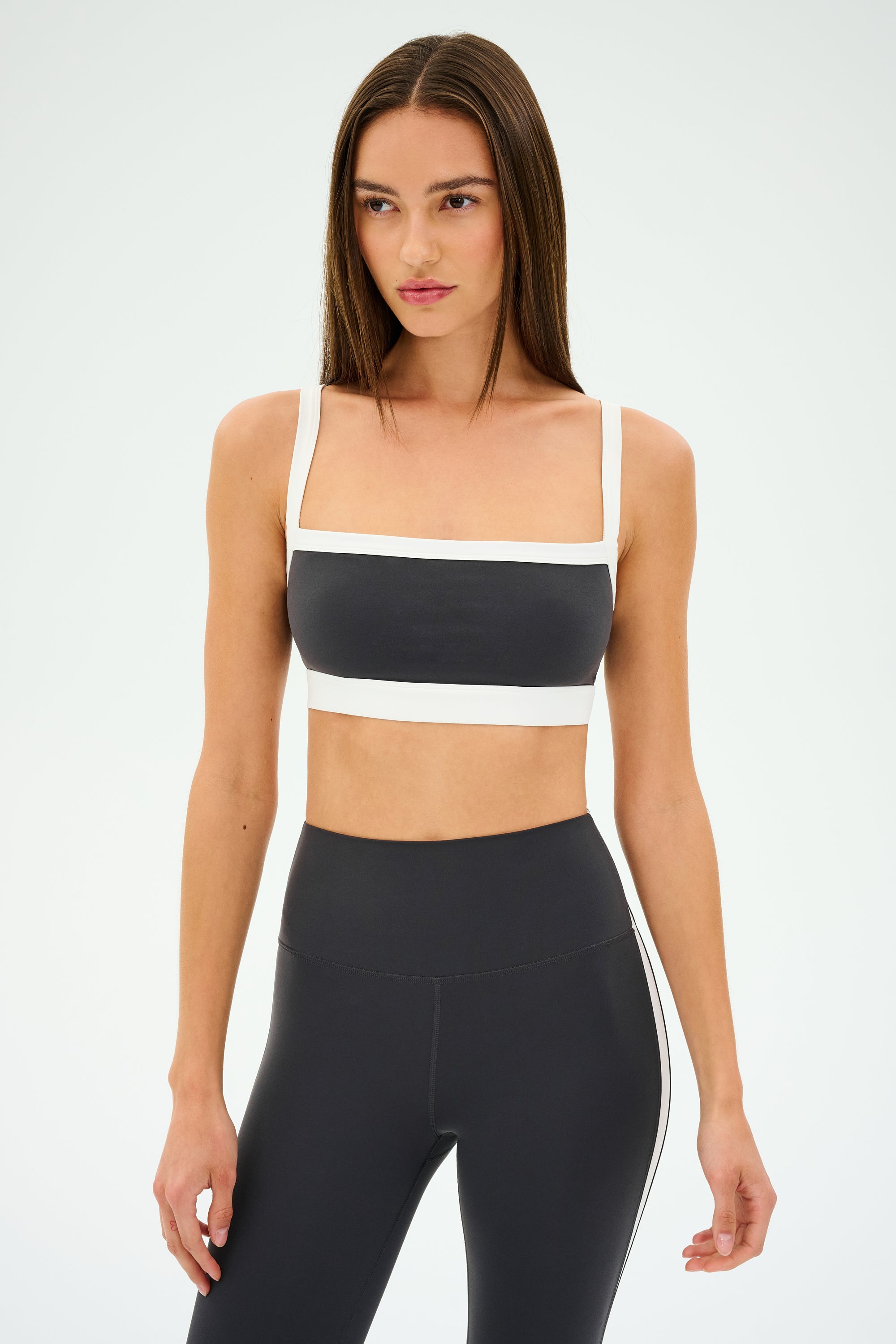 Poised against a plain backdrop, a person wearing a chic SPLITS59 Monah Rigor Bra in Graphite/White and black leggings is ready to conquer their gym workouts.