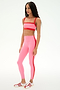 A woman stands in a studio wearing SPLITS59's Clare High Waist Rigor 7/8 leggings in Strawberry/Lotus Red, perfect for gym workouts, paired with red and white sneakers and a pink sports bra.