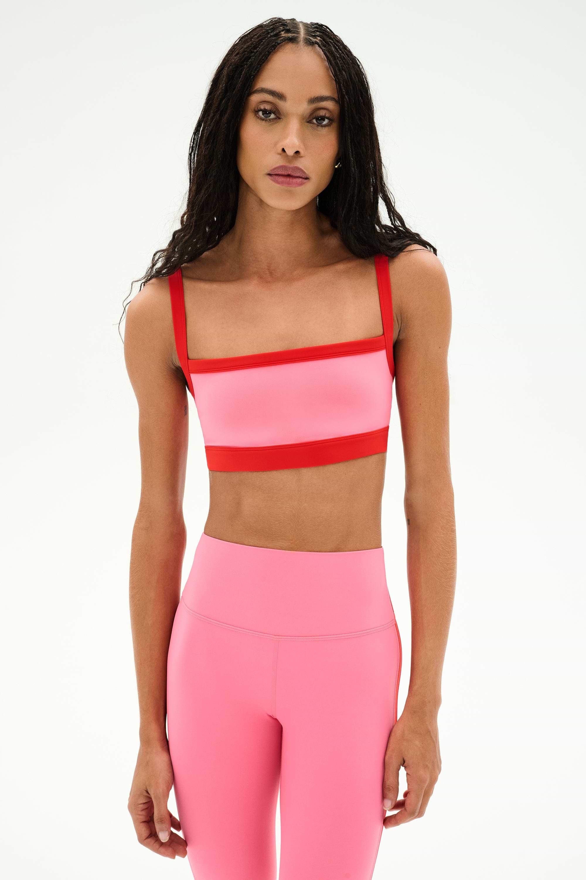 A woman wearing a SPLITS59 Monah Rigor Bra in Strawberry/Lotus Red and matching high-waisted leggings stands against a plain white background.