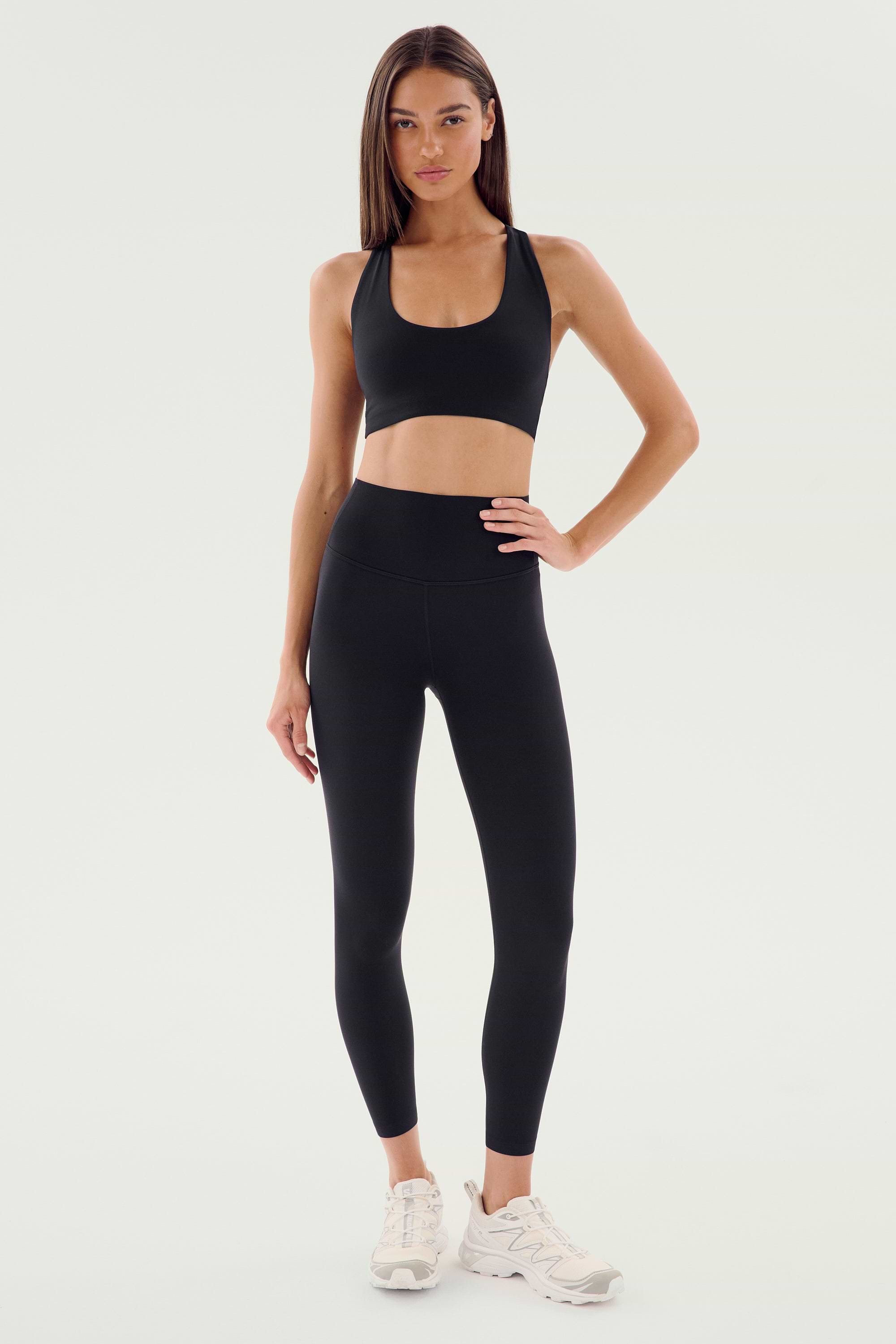 A woman stands against a plain background wearing the SPLITS59 Airweight High Waist Legging in black, paired with a black sports bra and white athletic shoes—an ideal look for hot yoga.