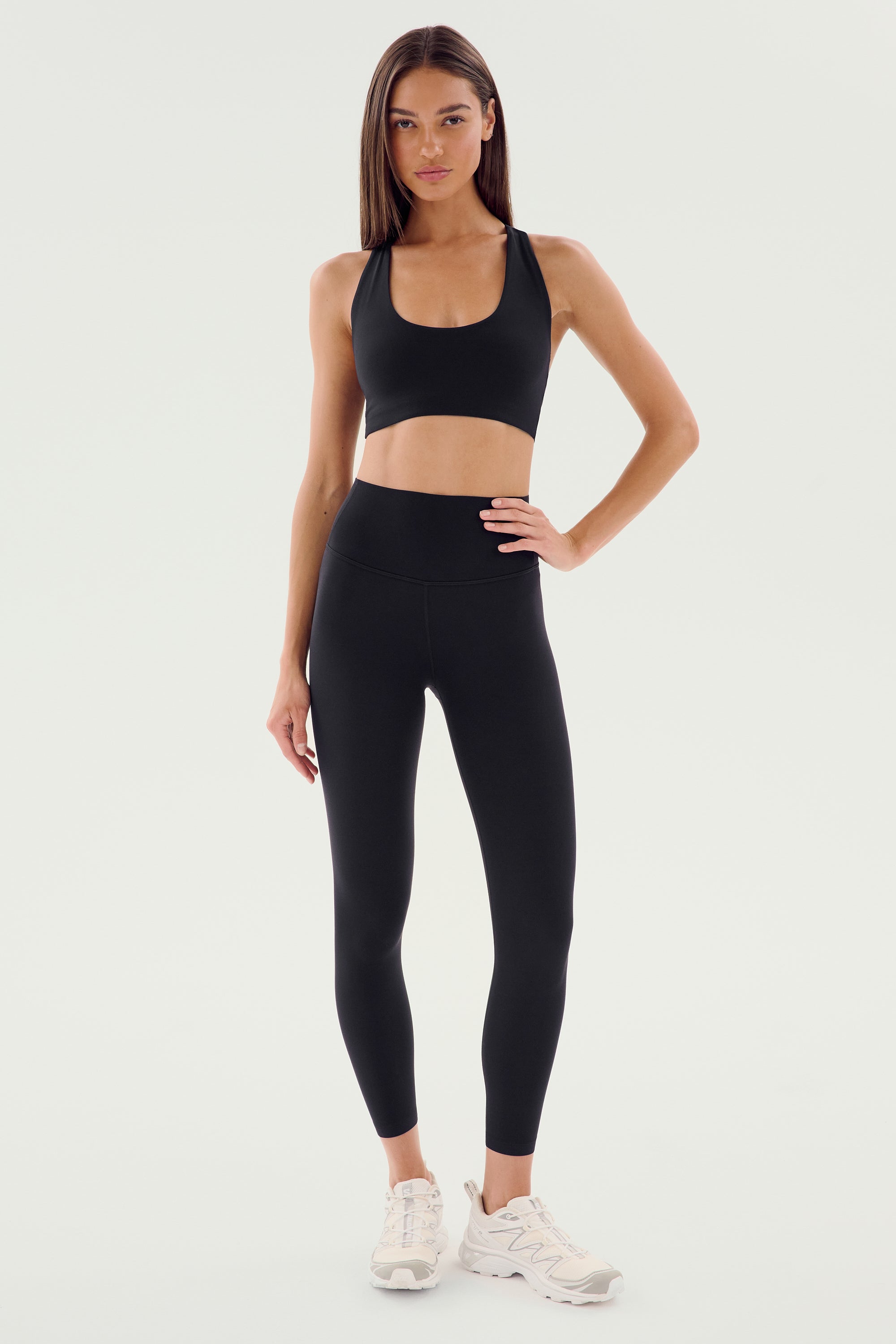 A woman stands against a plain background wearing the SPLITS59 Airweight High Waist Legging in black, paired with a black sports bra and white athletic shoes—an ideal look for hot yoga.