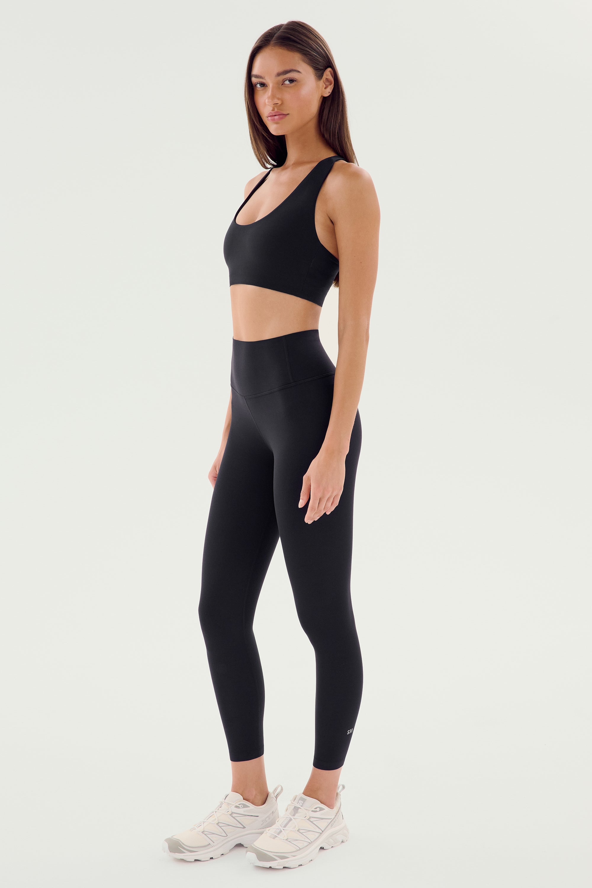 A woman wears SPLITS59’s Airweight High Waist Legging in black with a matching sports bra and white athletic shoes, standing against a plain white background.