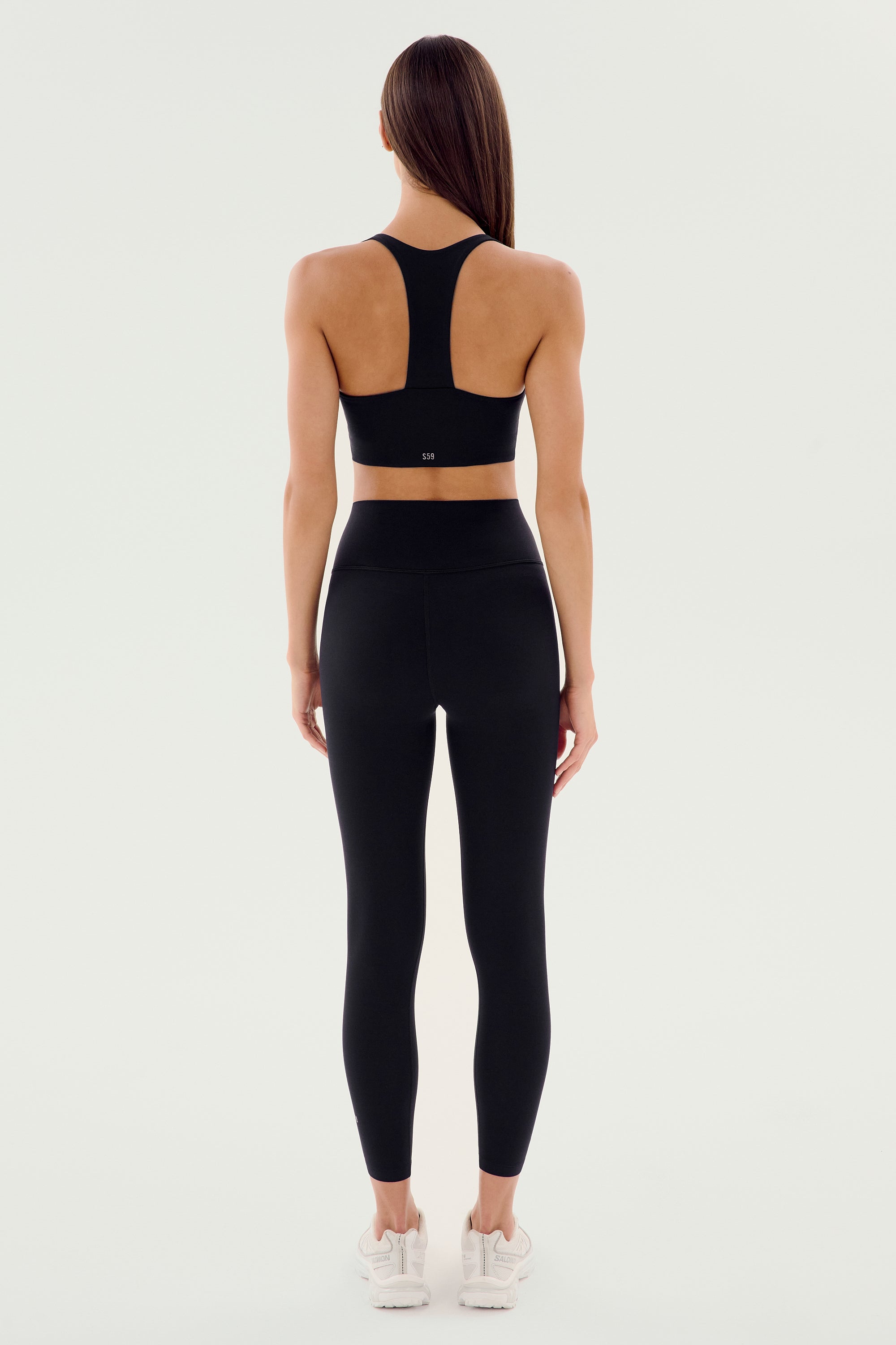 A woman stands with her back to the camera, wearing the SPLITS59 Airweight High Waist Legging in black—ideal for hot yoga—paired with a matching sports bra and white sneakers against a plain light background.