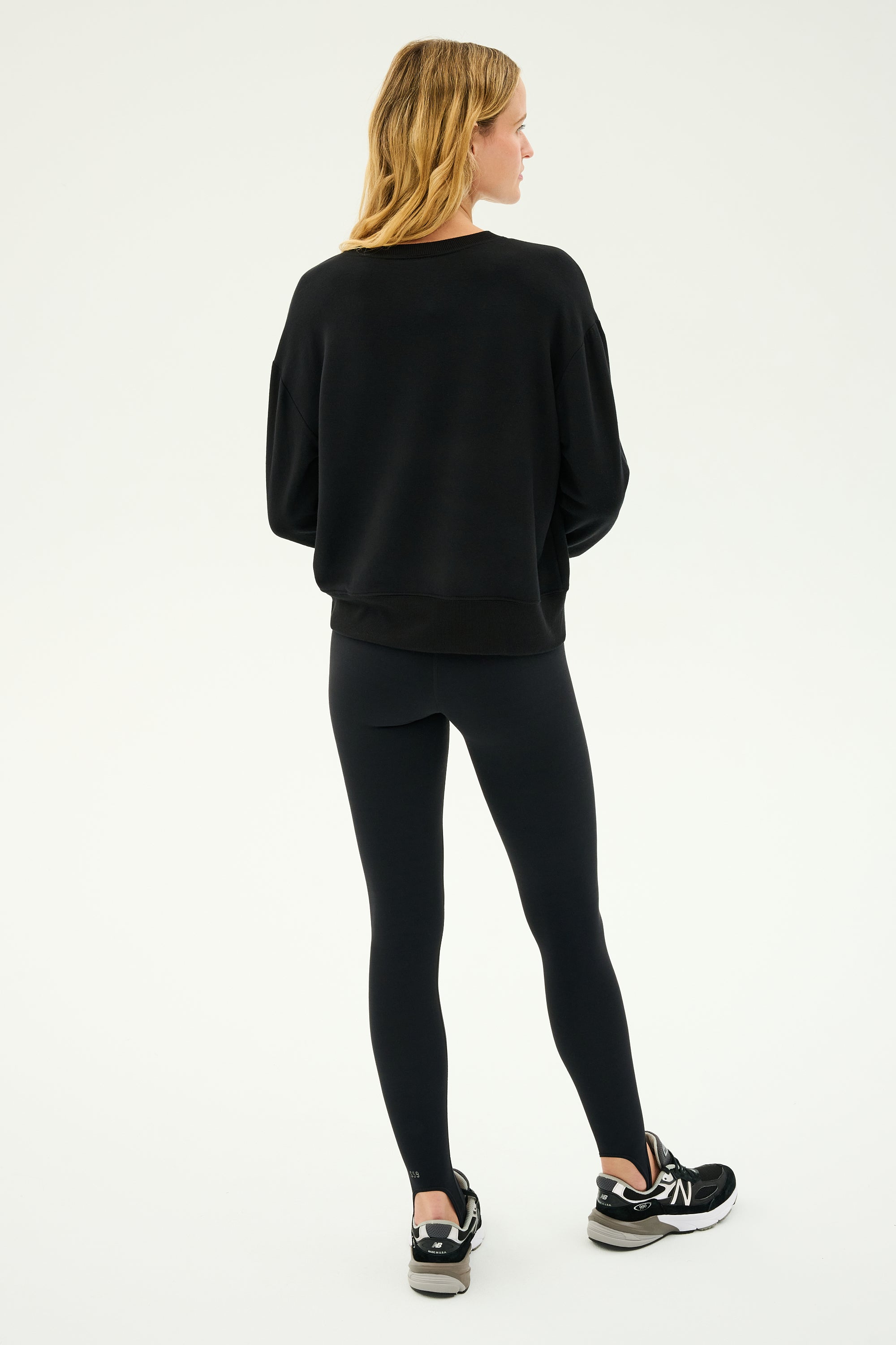 A person is standing against a neutral background, wearing the Sonja Fleece Sweatshirt in black crafted from ultra-luxe fleece by SPLITS59, paired with leggings and sneakers. The outfit effortlessly embodies comfort and style, exemplifying the MADE IN LOS ANGELES quality.