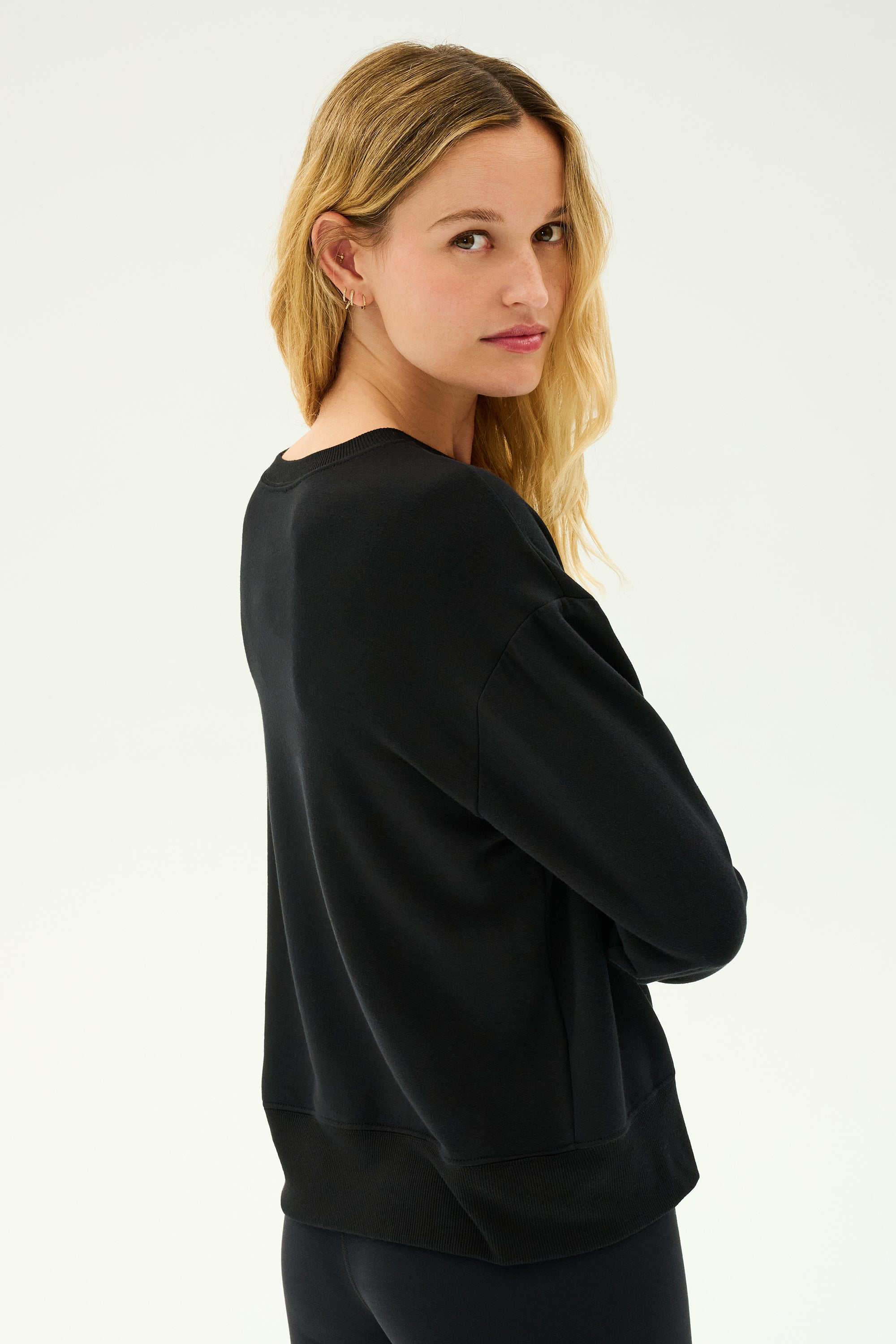 A woman with long blonde hair is wearing the SPLITS59 Sonja Fleece Sweatshirt in black paired with matching pants, standing against a light background and looking over her shoulder.