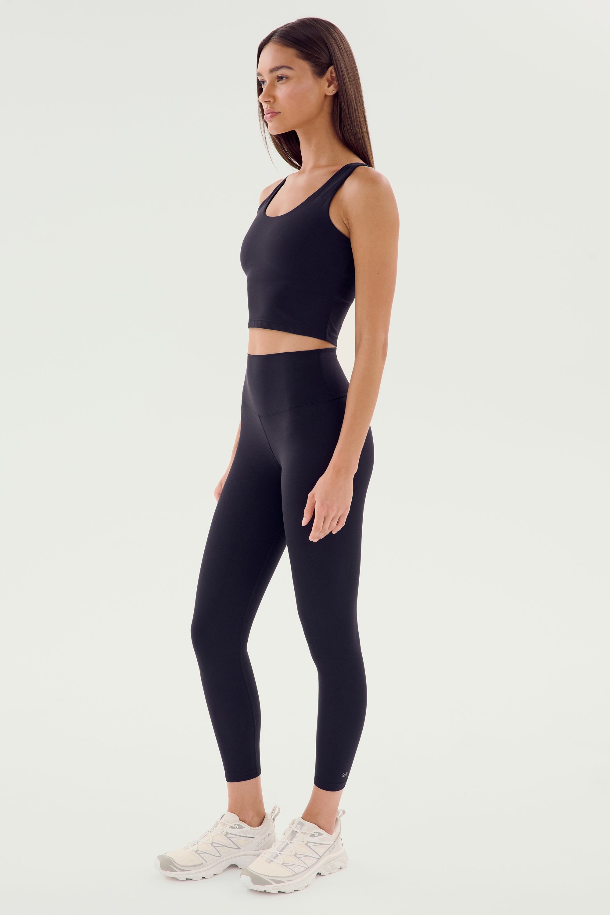 A person stands sideways wearing a black Sprint High Waist Rigor 7/8 from SPLITS59, a matching black sports bra, and white sneakers against a plain background.