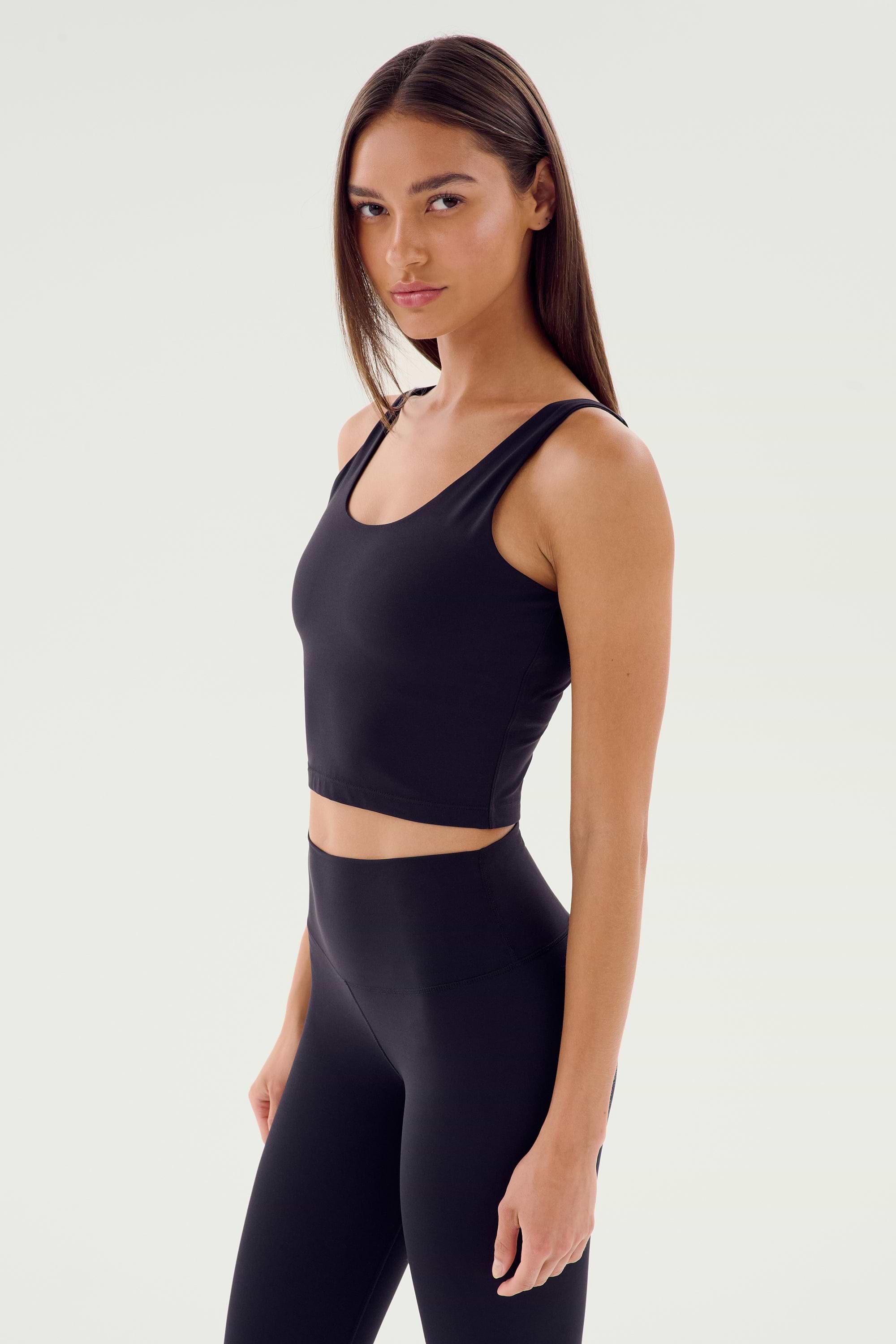 A person with long hair wearing a black Sprint Rigor Bralette by SPLITS59 and high-waisted leggings stands against a plain background.