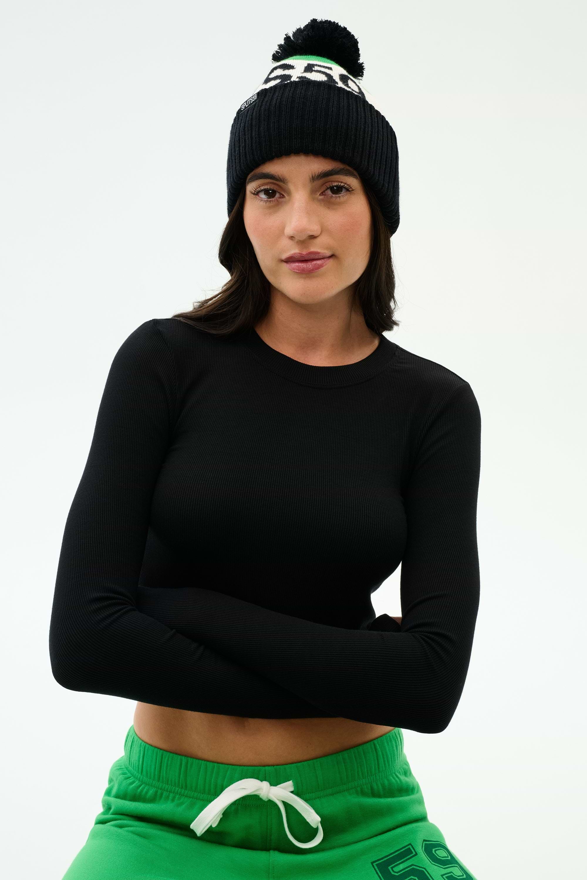 Woman wearing a black long-sleeve top, green drawstring pants, and a black beanie with a pom-pom, standing with arms crossed against a plain background.