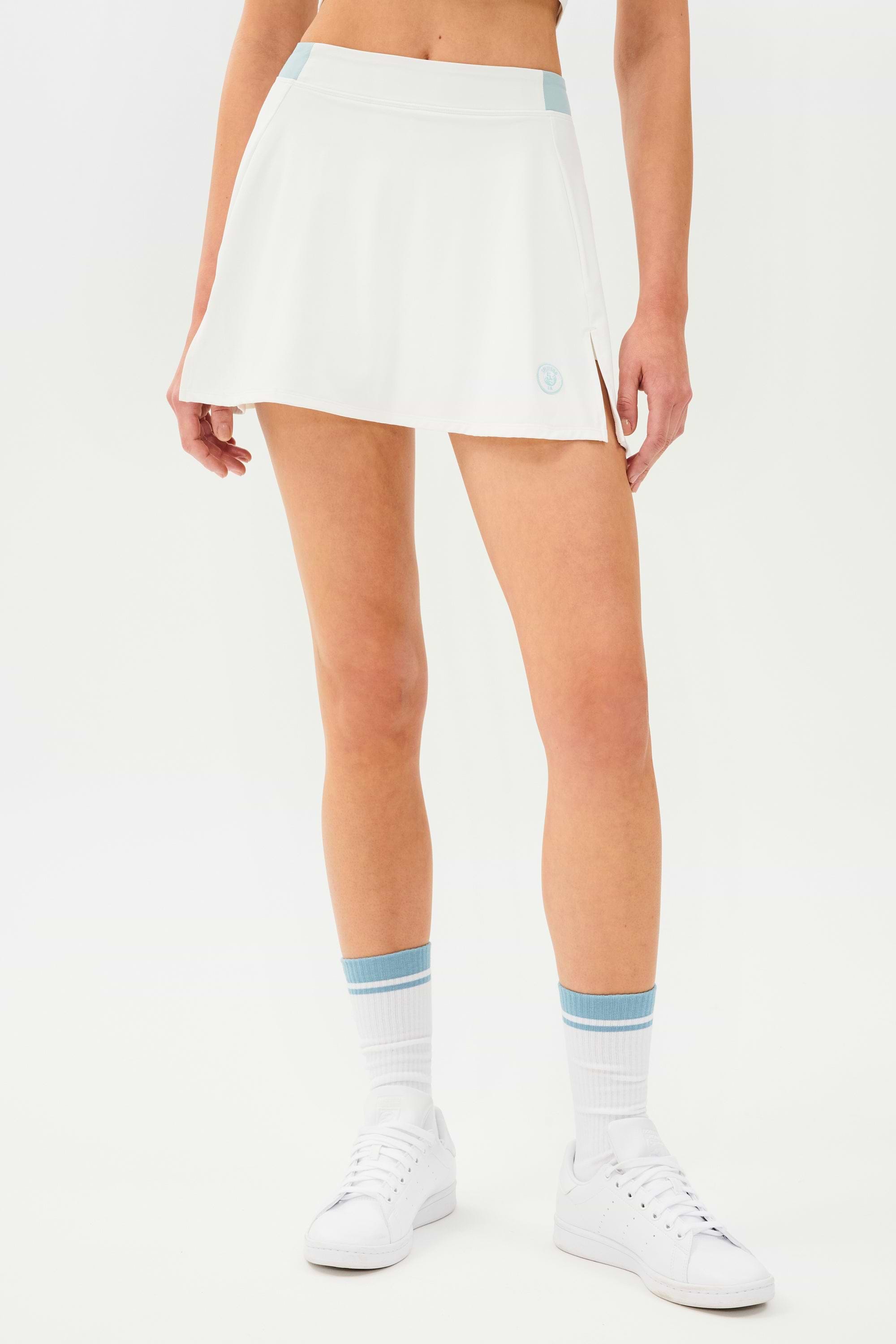 Front full view of white skort with teal details on the waistband and logo. Model is wearing white socks with teal stripe and all white shoes