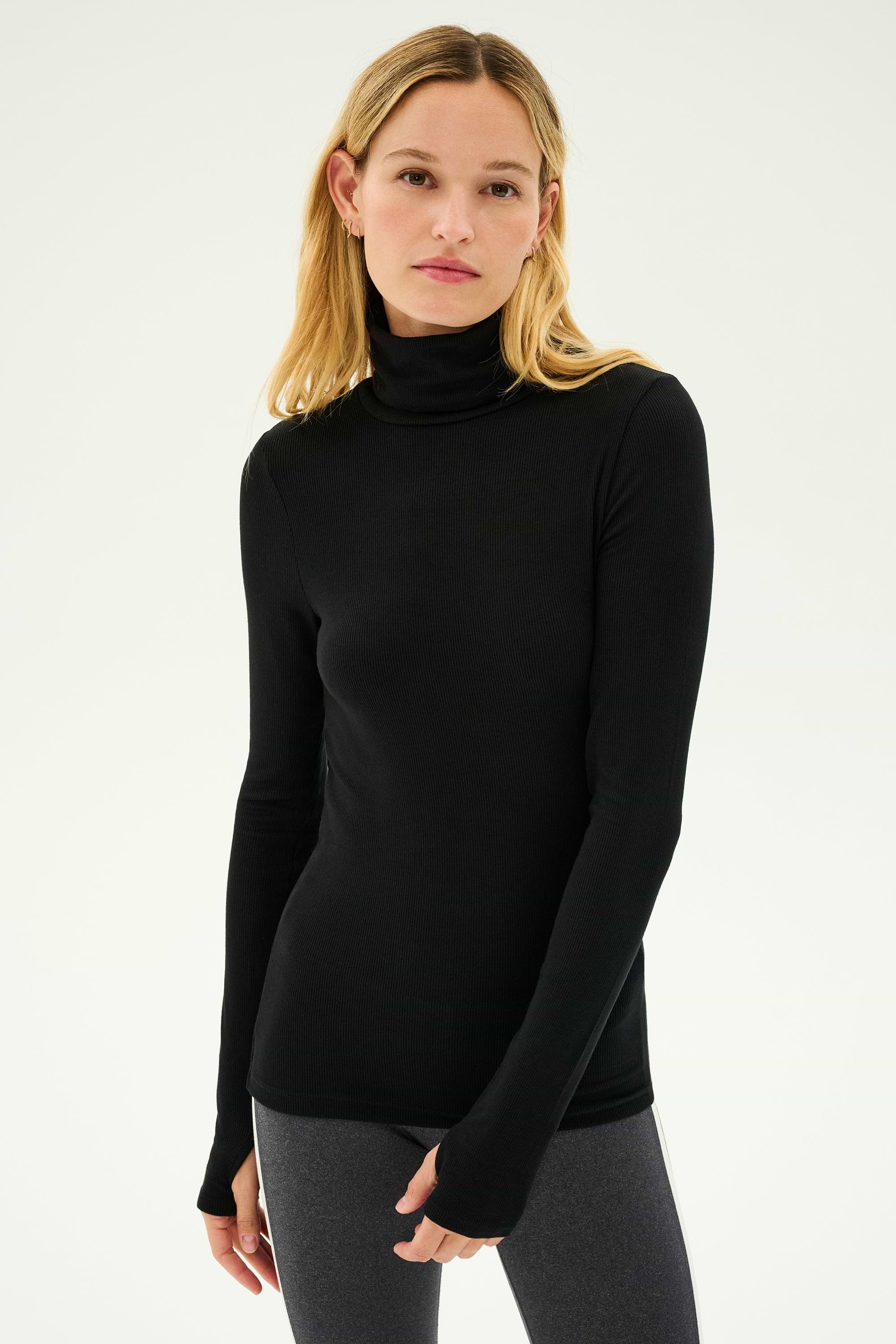 A person with long, light-colored hair is wearing a black Jackson Rib Full Length Turtleneck from SPLITS59 and gray bottoms, standing against a plain white background.