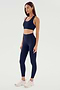 Person wearing a matching indigo Sara Airweight Bra from SPLITS59 and leggings, standing confidently with arms relaxed by their sides and looking to the left. White sneakers complete the athletic outfit, perfect for Pilates or hot yoga sessions.