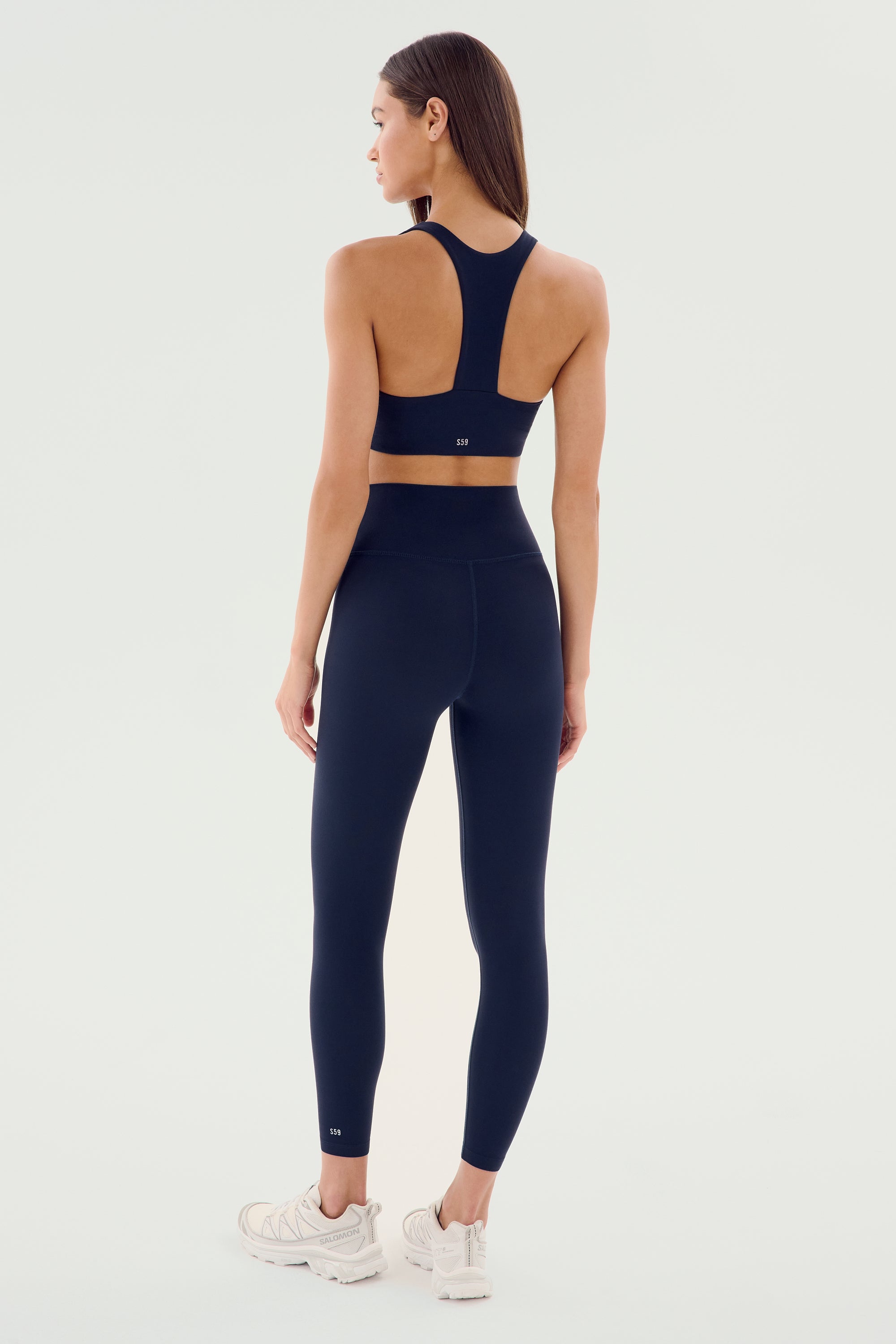 A woman stands with her back to the camera wearing the SPLITS59 Airweight High Waist Legging in Indigo and a matching navy blue sports bra, complemented by white sneakers, against a plain background.