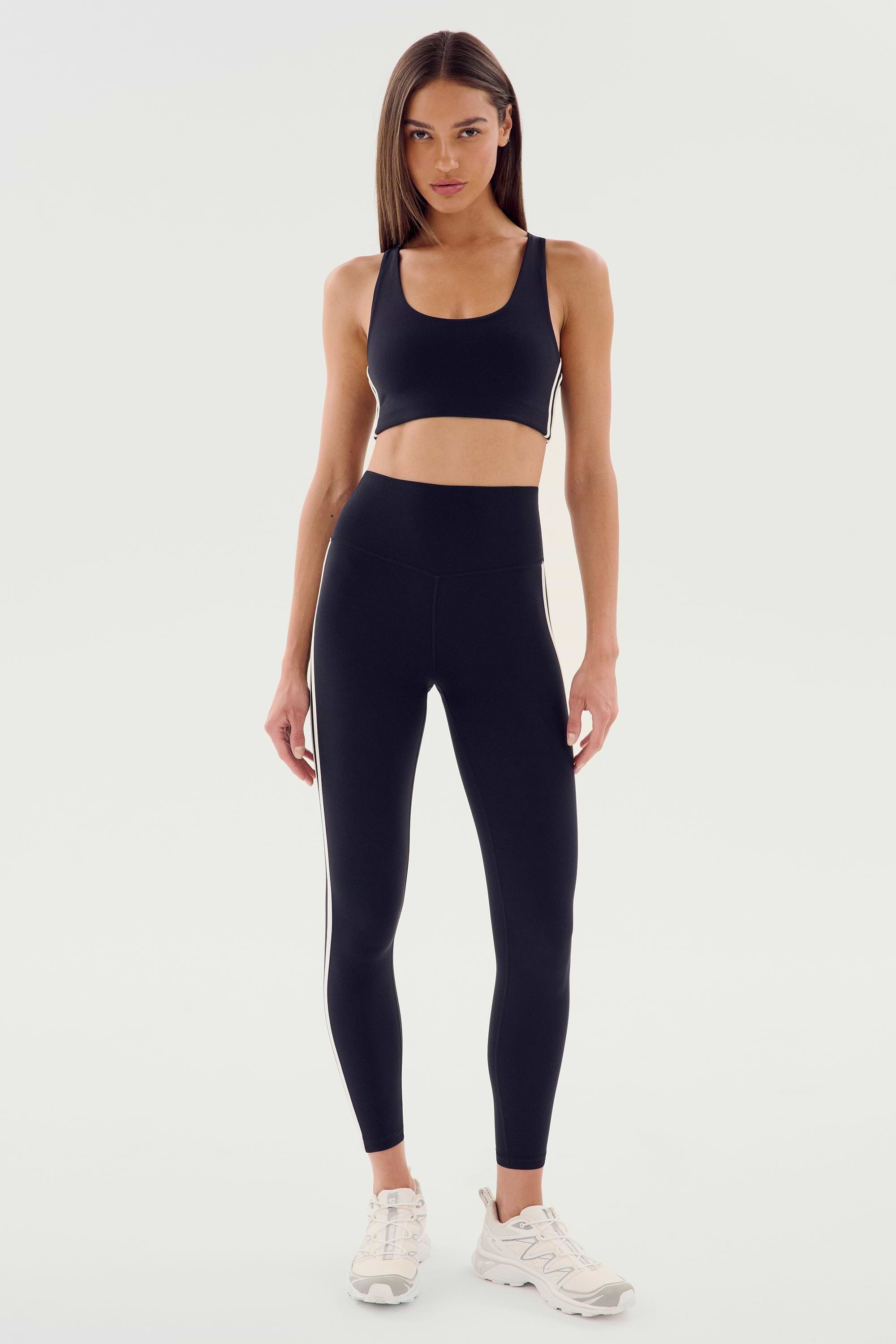 A person standing and wearing the SPLITS59 Ella Airweight Bra in Black/White paired with matching leggings featuring white side stripes and white sneakers, set against a plain background.