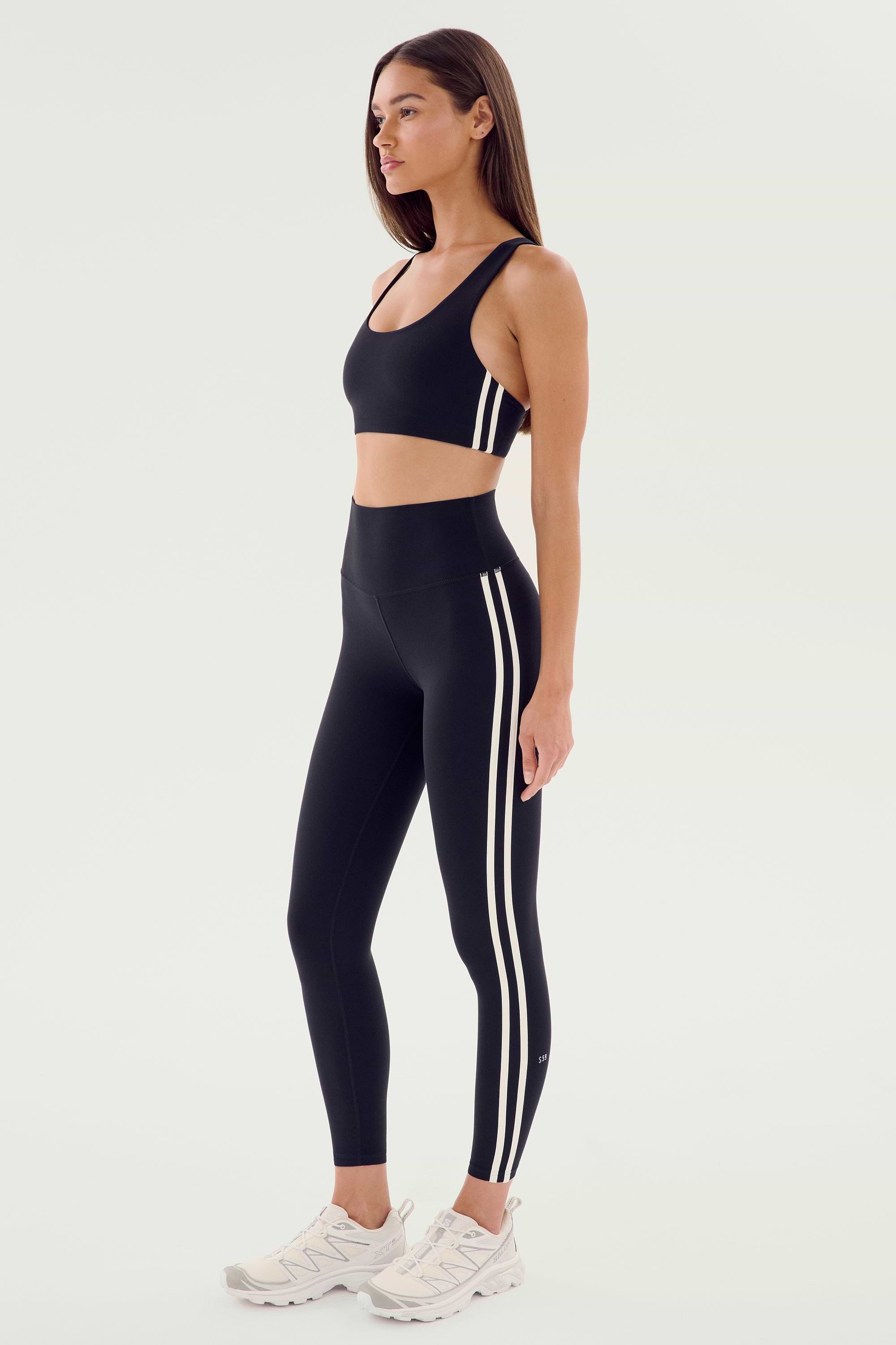 A woman wearing the SPLITS59 Ella Airweight Bra in black and white, along with black leggings featuring white stripes, stands in a side profile against a plain background. She completes her look with white sneakers.
