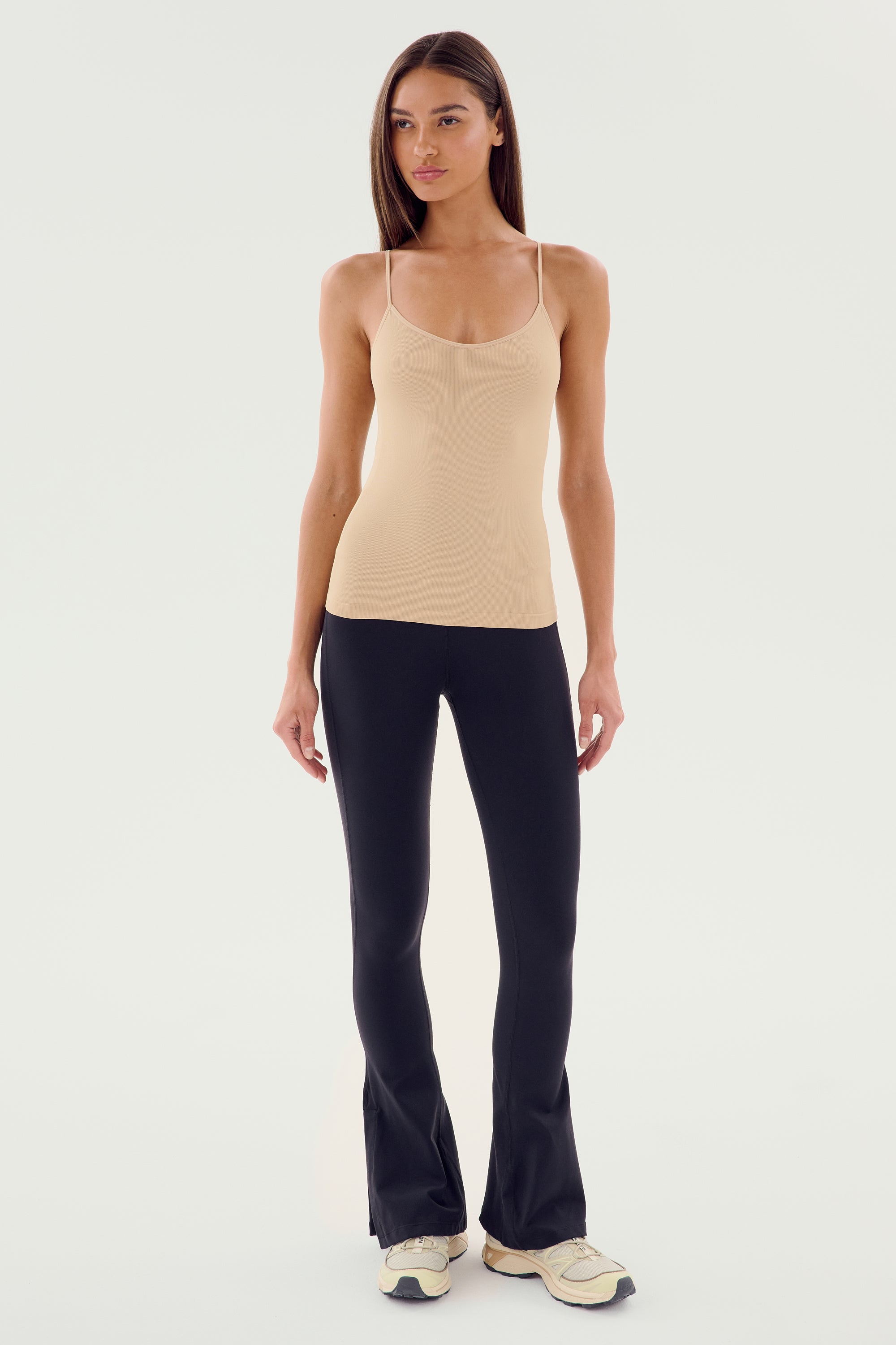 A person stands against a plain background wearing the Loren Seamless Waist Length Tank in Nude by SPLITS59, paired with black pants and white sneakers—perfect for gym workouts.