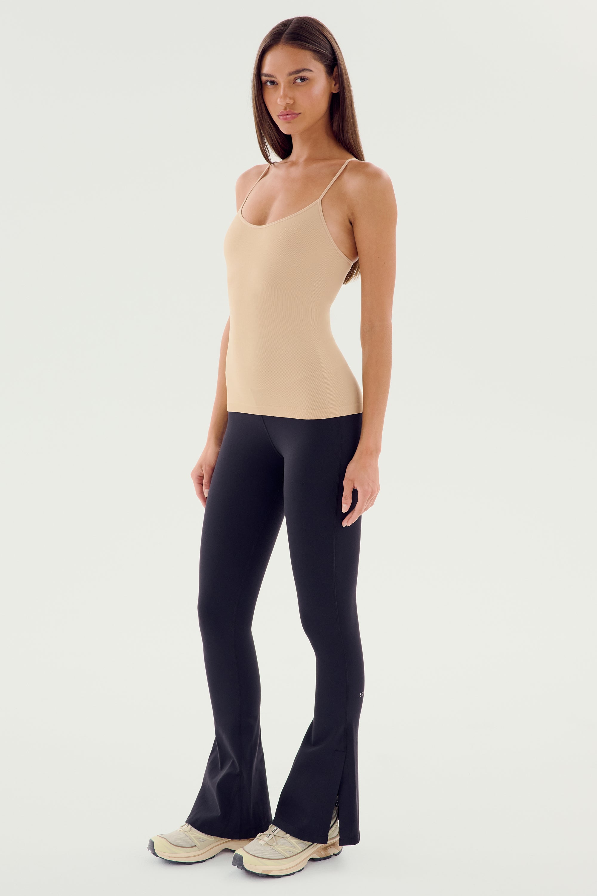 A woman stands against a plain background wearing a beige tank top, SPLITS59's Raquel High Waist Flare w/ Split Hem pants in black, and white sneakers.