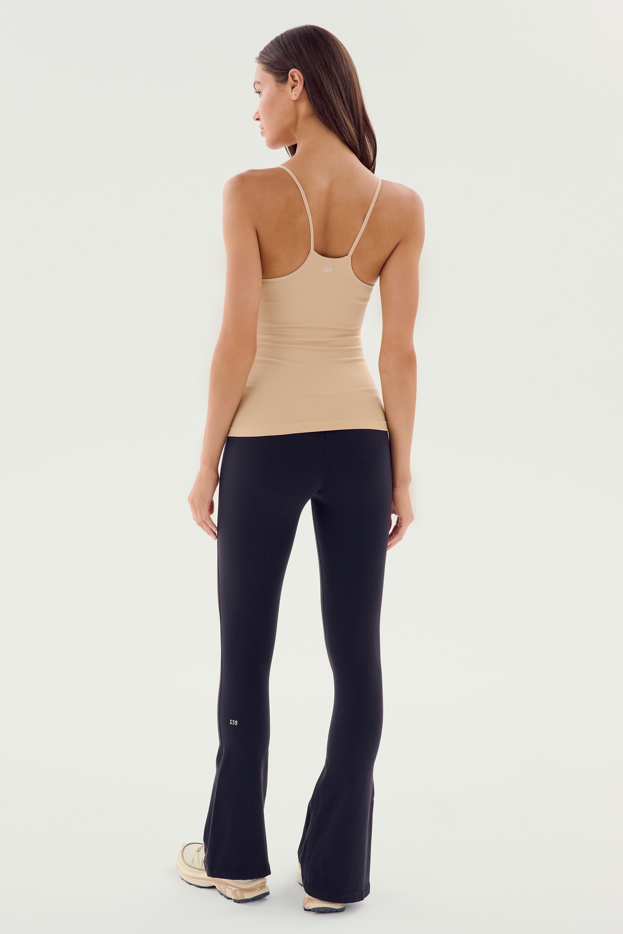 A woman stands facing away, her hair down, wearing a beige sleeveless top and the SPLITS59 Raquel High Waist Flare w/ Split Hem - Black made from 4-way stretch supplex fabric. She has light-colored sneakers and is standing against a plain light background.