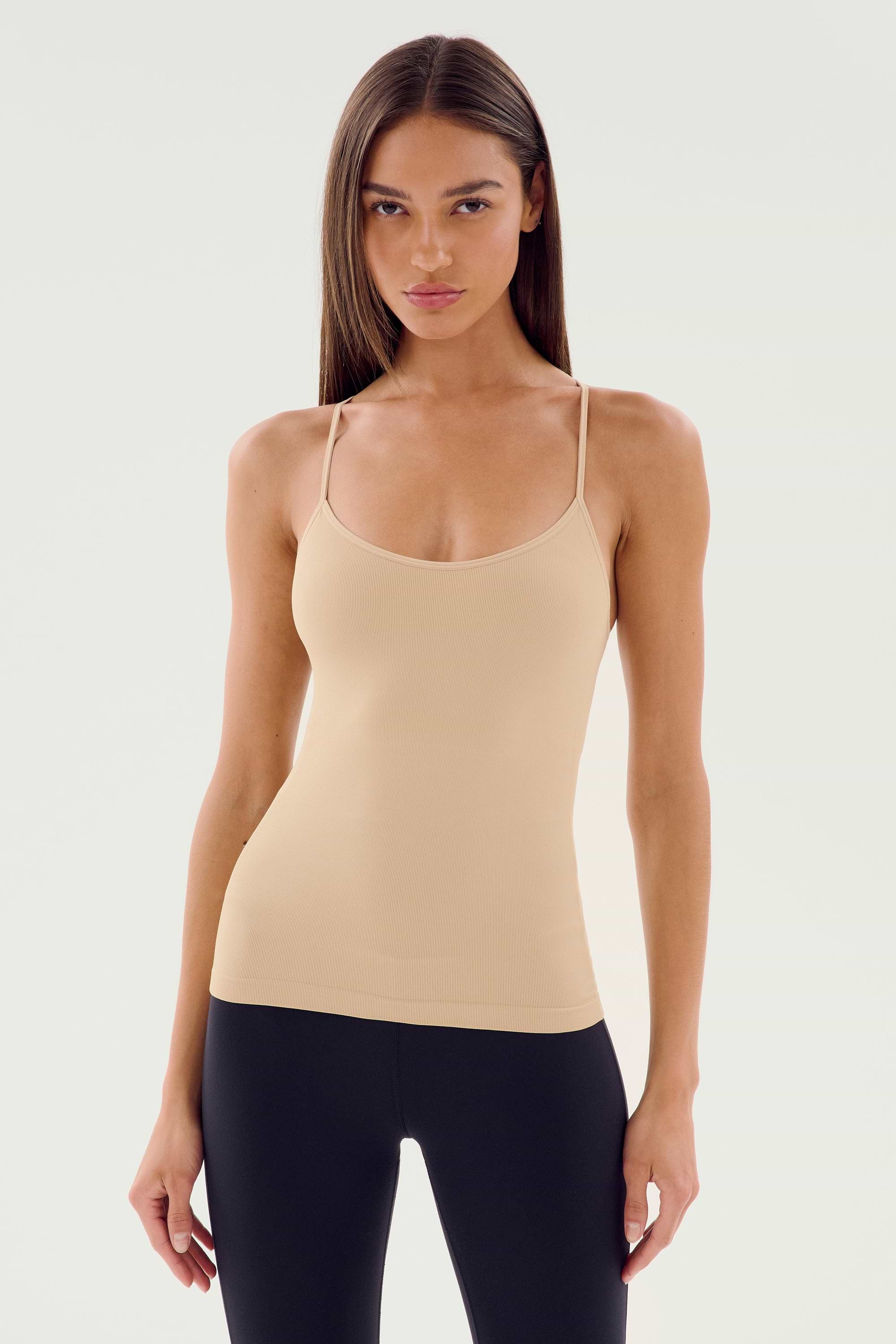 A person with long hair wearing the SPLITS59 Loren Seamless Waist Length Tank in Nude and black pants stands against a plain background.