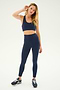 A person stands against a plain background, wearing a SPLITS59 navy sports bra and the Airweight Super High Waist 7/8 leggings in Indigo by the same brand, paired with black athletic shoes. They have one hand behind their head and the other hand by their side, ready for hot yoga.