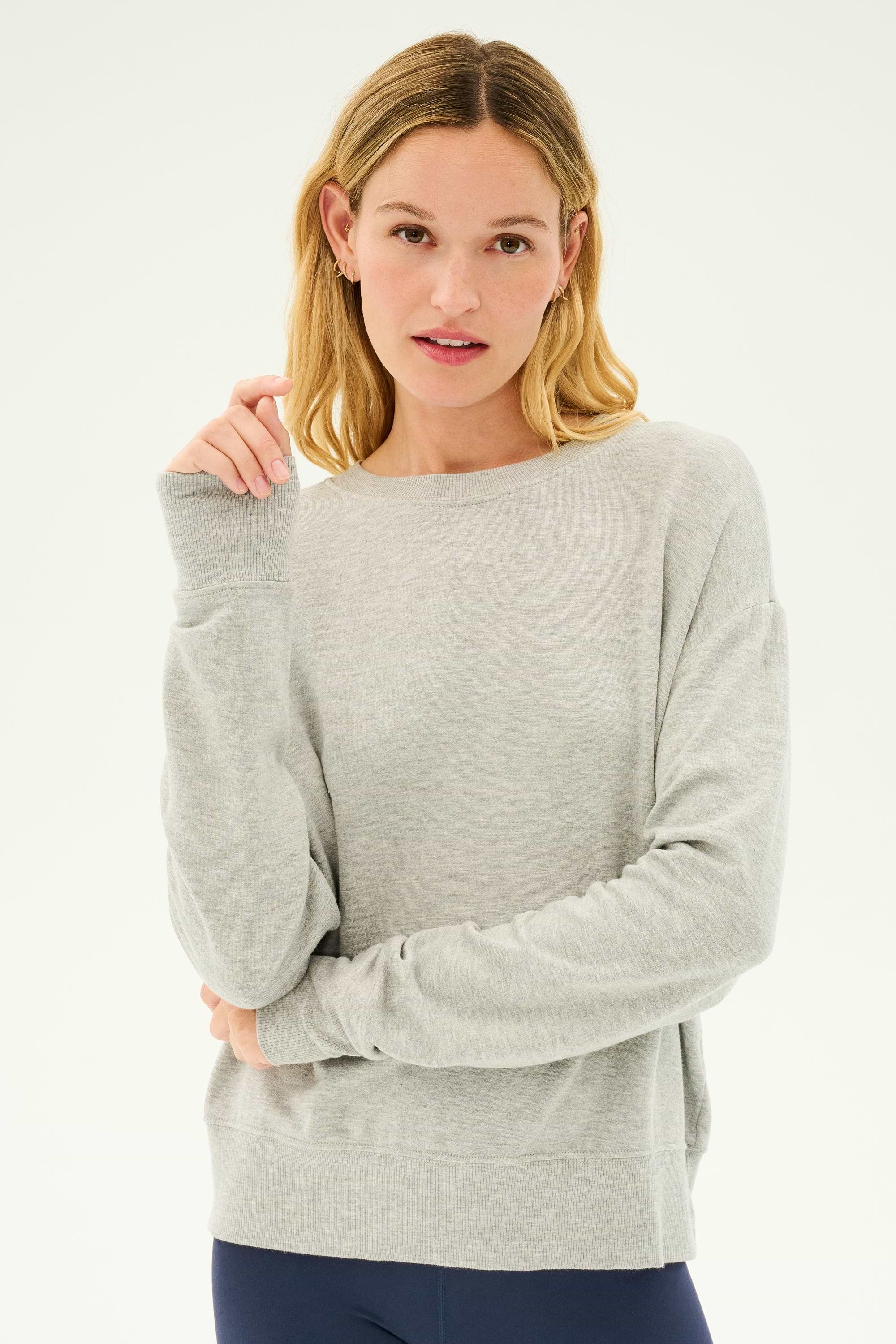 A person with blonde hair wearing the Sonja Fleece Sweatshirt in Heather Grey by SPLITS59 and dark pants, made in Los Angeles with ultra luxe fleece, stands against a plain background.