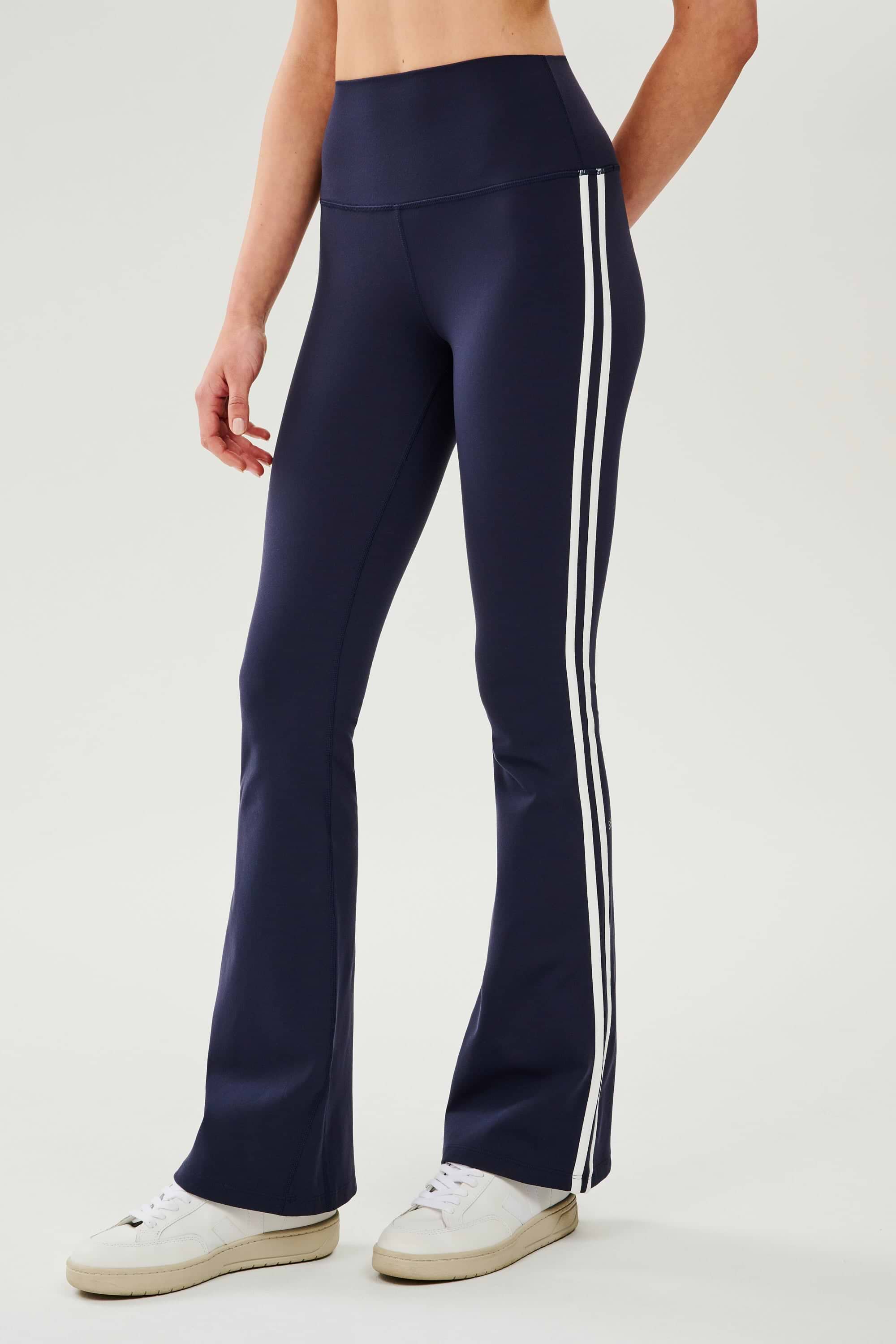 Front view of woman wearing dark blue high waist below ankle length legging with wide flared bottoms and white double side stripes on both legs. Paired with white shoes.