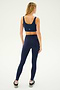 A person stands facing away, showcasing workout fitness attire in a navy blue sports bra, Sprint High Waist Rigor 7/8 leggings - Indigo by SPLITS59, and black sneakers on a light background.