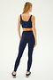 A person stands facing away, showcasing workout fitness attire in a navy blue sports bra, Sprint High Waist Rigor 7/8 leggings - Indigo by SPLITS59, and black sneakers on a light background.