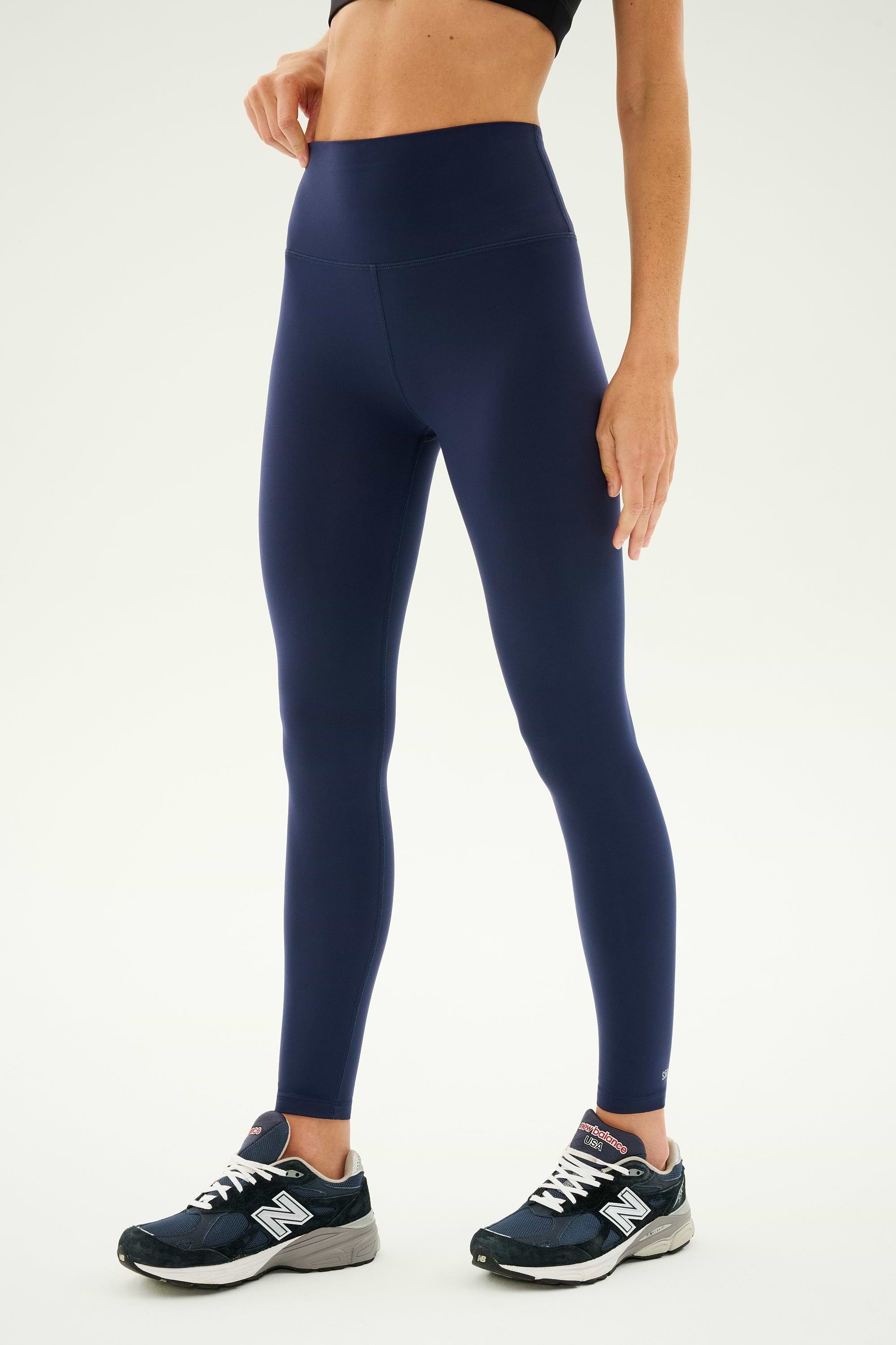 A person wearing a black sports bra, Sprint High Waist Rigor 7/8 leggings in Indigo from SPLITS59, and black running shoes stands against a plain white background.