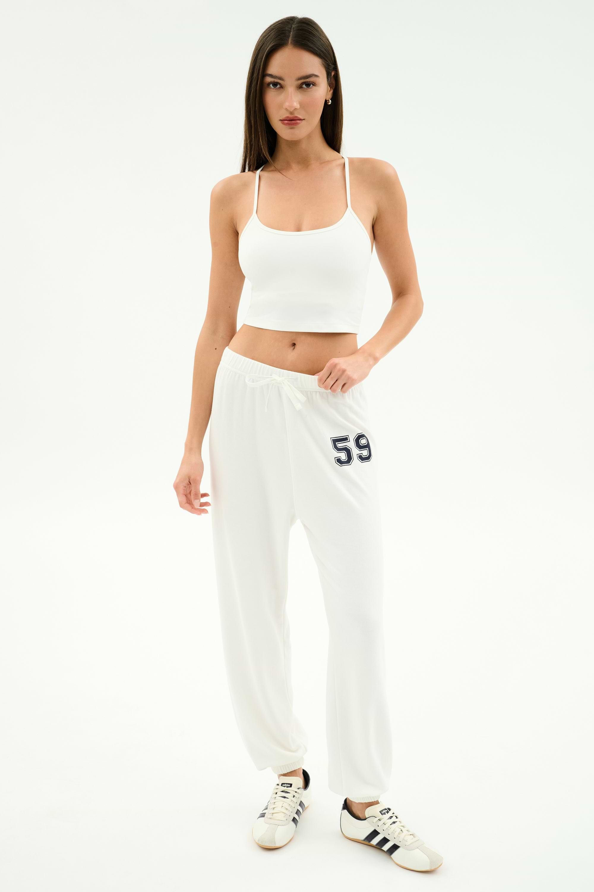 Woman standing against a white background wears a white crop top, SPLITS59 Andie Oversized Fleece Varsity 59 Sweatpant in White/59, and white Adidas sneakers—ideal for cool weather workouts. MADE IN LOS ANGELES.