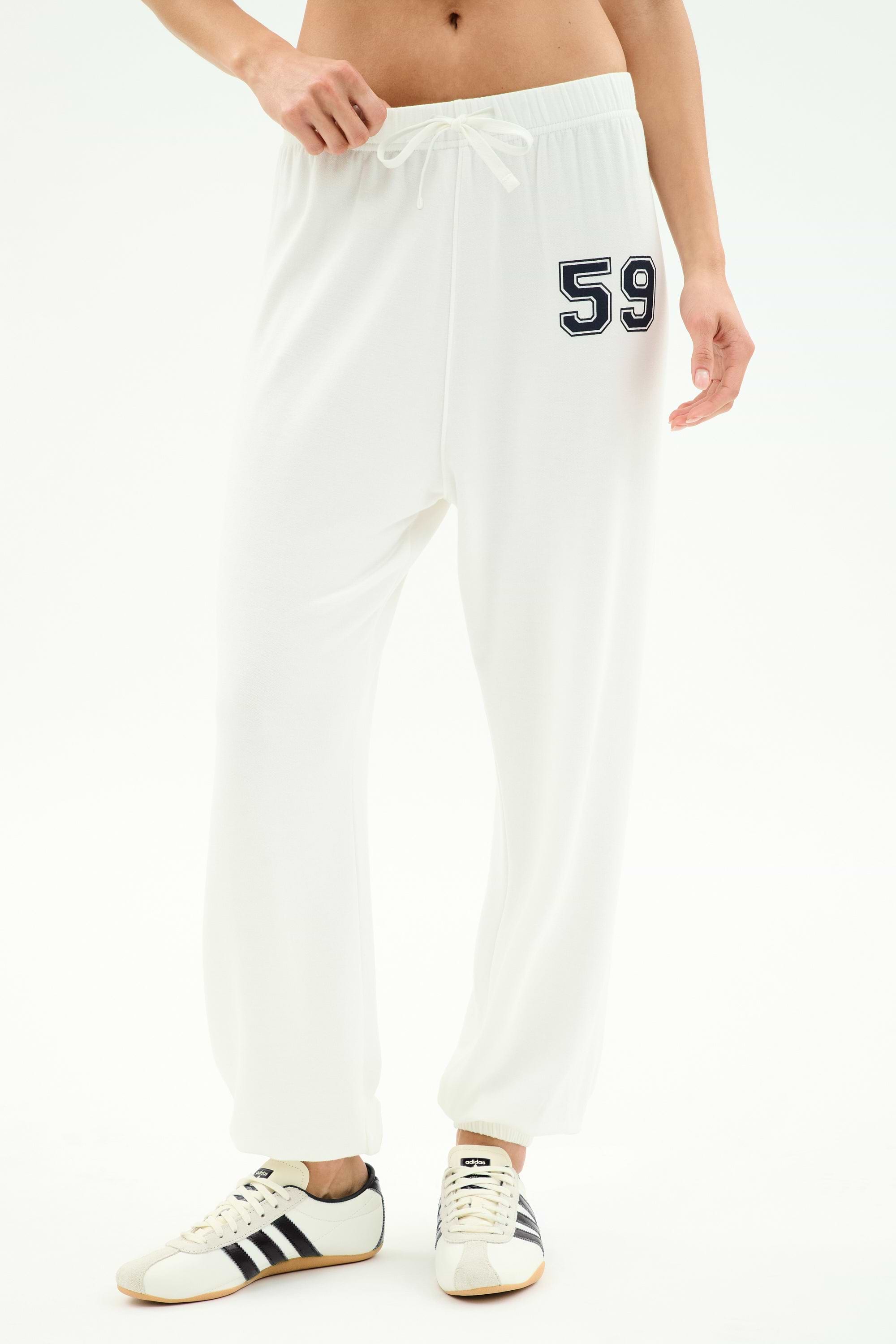 A person wears SPLITS59's Andie Oversized Fleece Varsity 59 Sweatpant in White/59, featuring the number 59 on the left hip, paired with white sneakers with black stripes—ideal for cool weather workouts—against a plain white background.