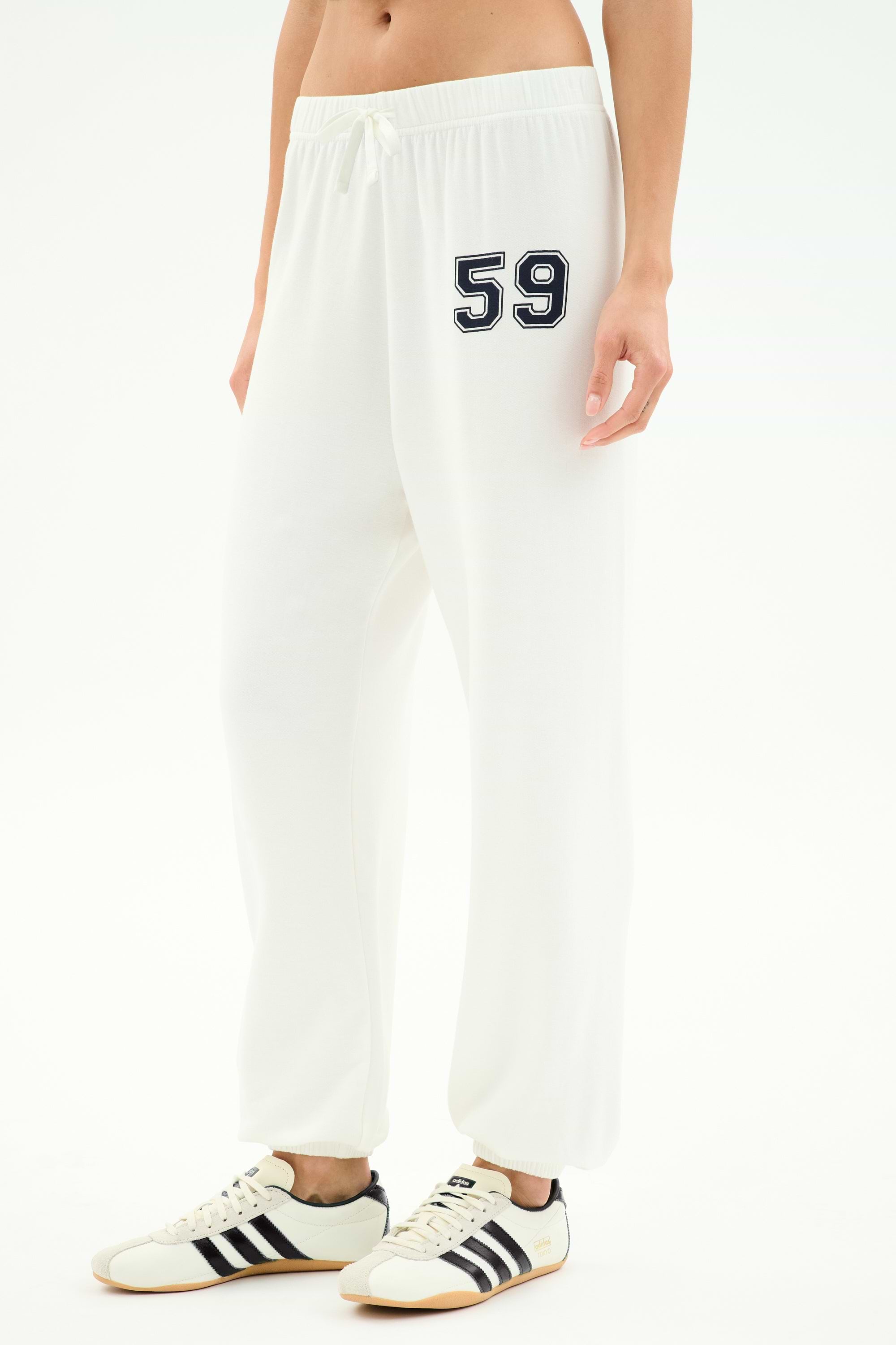 A person in SPLITS59 Andie Oversized Fleece Varsity 59 Sweatpant—white with bold "59"—paired with white sneakers with black stripes, perfect for cool weather workouts. Made in Los Angeles.